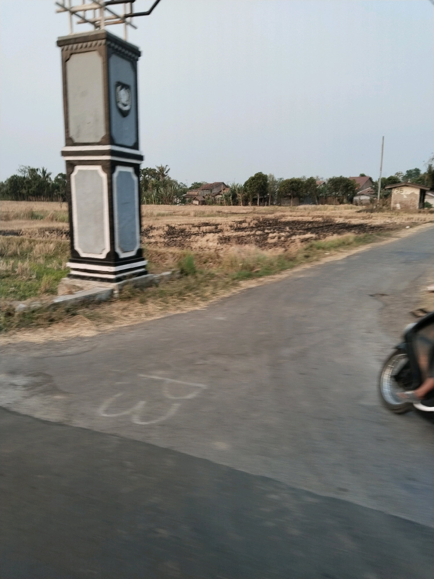 In Semboja, Indonesia, a street scene unfolds with a stone monument, a dry field, and a moving motorcycle. The monument, a multi-tiered structure with dark trim and light-colored panels, stands prominently on the left side of the frame. A patch of dry, straw-like vegetation is visible behind it, suggesting a rural or semi-rural environment. Further in the background, a cluster of houses and trees dots the landscape under a pale sky. The road in the foreground is paved, and on the right side, the rear wheel and part of the rider's leg on a motorcycle are visible, indicating movement. White chalk marks are faintly present on the road surface. The overall impression is one of a quiet, possibly agricultural area with ongoing daily activity.