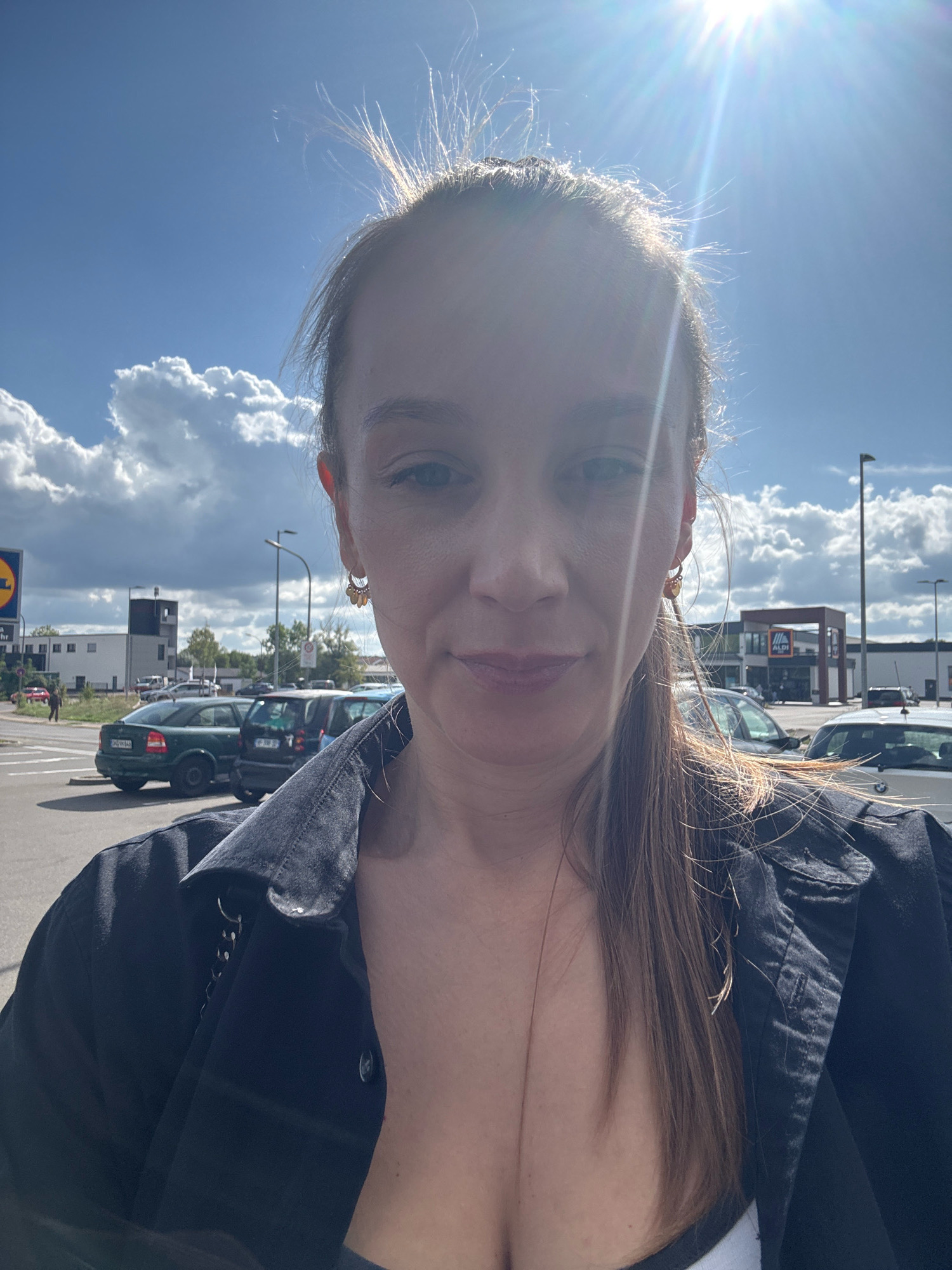 A woman is taking a selfie outdoors in a parking lot in front of an ALDI store in Überherrn, Germany. The sun is bright and shining, creating lens flare in the upper portion of the image. The woman is wearing a dark jacket and has long brown hair. In the background, there are cars parked in the lot and the ALDI store is visible. There are also clouds in the sky.