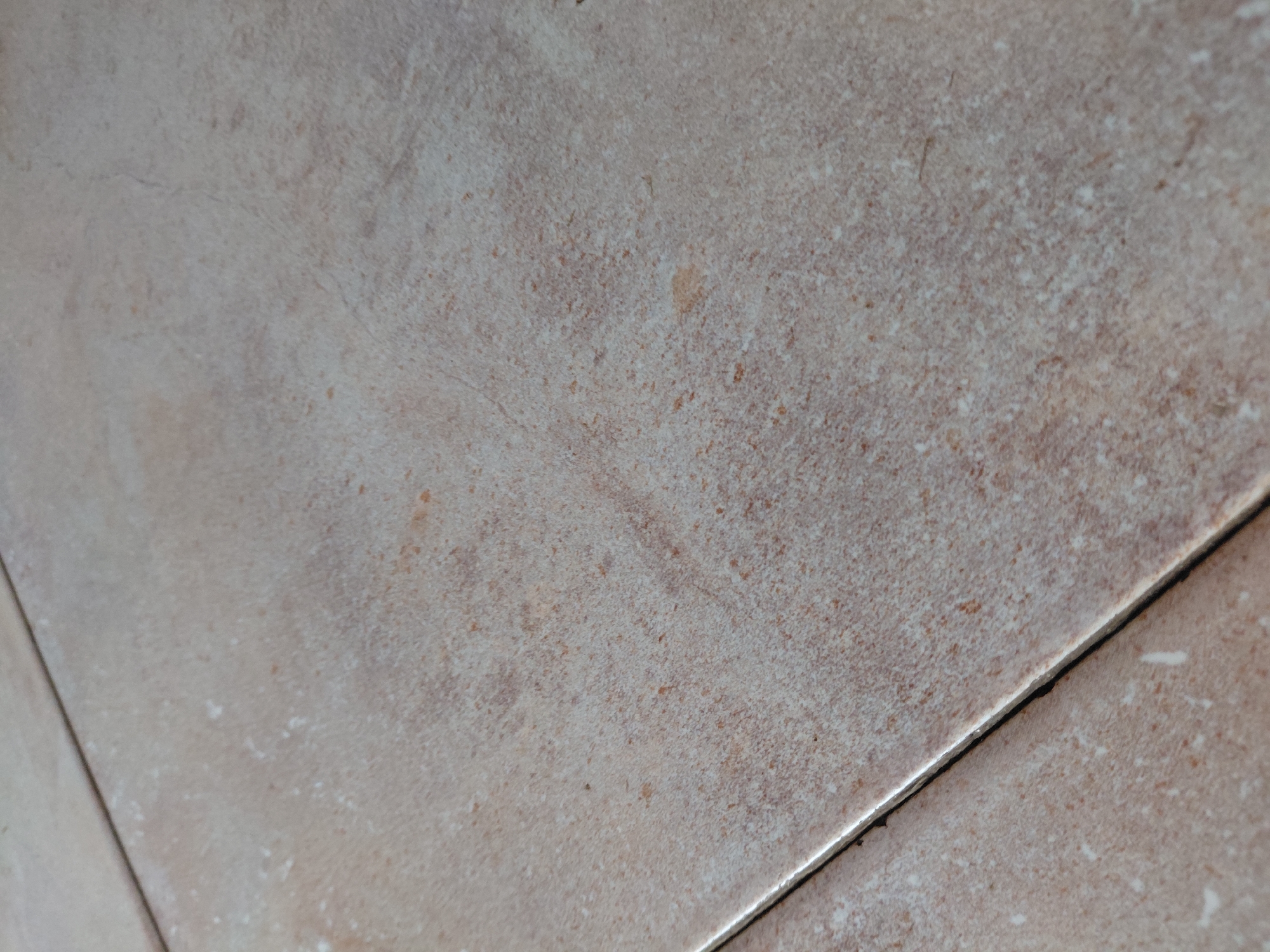 The image presents a close-up, angled view of a tiled surface, likely a floor, in Santa Ana, Venezuela. No people, objects, or visible text are present.
The tiles are predominantly light brown and beige, featuring a mottled, textured surface that resembles natural stone. Their irregular pattern includes scattered flecks and patches of darker brown, reddish-brown, and lighter off-white tones, contributing to a variegated and rustic aesthetic. The surface appears to have a matte or semi-matte finish.
Two dark grout lines are visible, running diagonally across the lower and central parts of the frame, meeting at an acute angle in the lower right quadrant. The grout is dark grey or black, contrasting with the lighter tiles, and shows slight discoloration or accumulation of dirt within its recessed channels. The edges of the tiles where they meet the grout appear clean. The lighting is soft and even, highlighting the texture without harsh shadows or bright reflections.