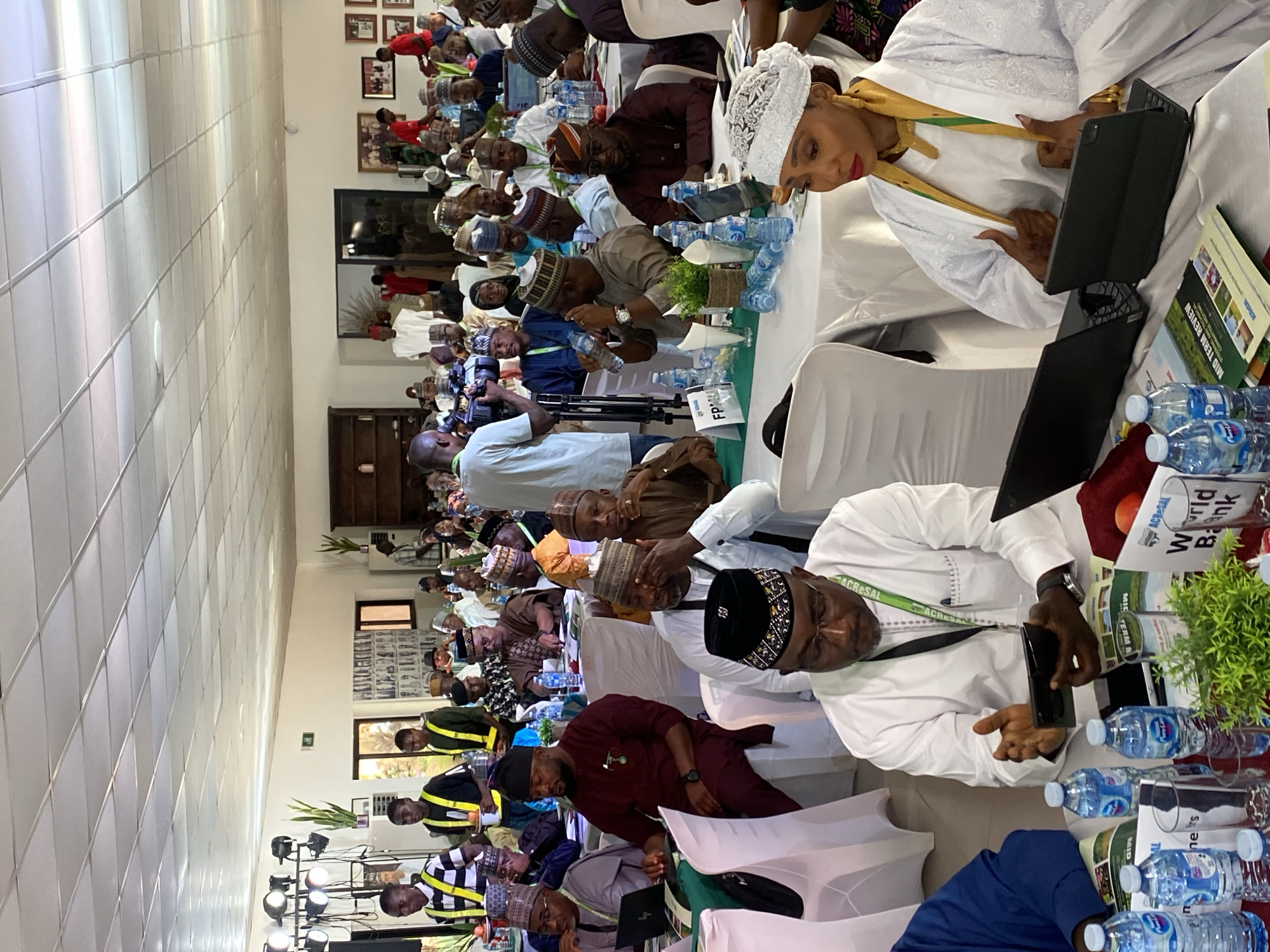 A photograph depicts a meeting or conference taking place indoors in what appears to be Kwarau, Nigeria. The room is populated by many people, mostly men, seated at tables covered with white tablecloths. The majority of the attendees are wearing traditional Nigerian attire, including hats, robes, and gowns. Several individuals have laptops and water bottles in front of them, and a few are engaged in using their mobile phones or tablets.
The backdrop includes photographs on the wall, indicating a formal setting. In the foreground, there are multiple water bottles placed on the table. The lighting in the room is bright, suggesting an indoor setting. The individuals present seem to be participating in a professional event, likely related to agricultural matters indicated by documents with the words "Mid Term Review," and "World Bank."