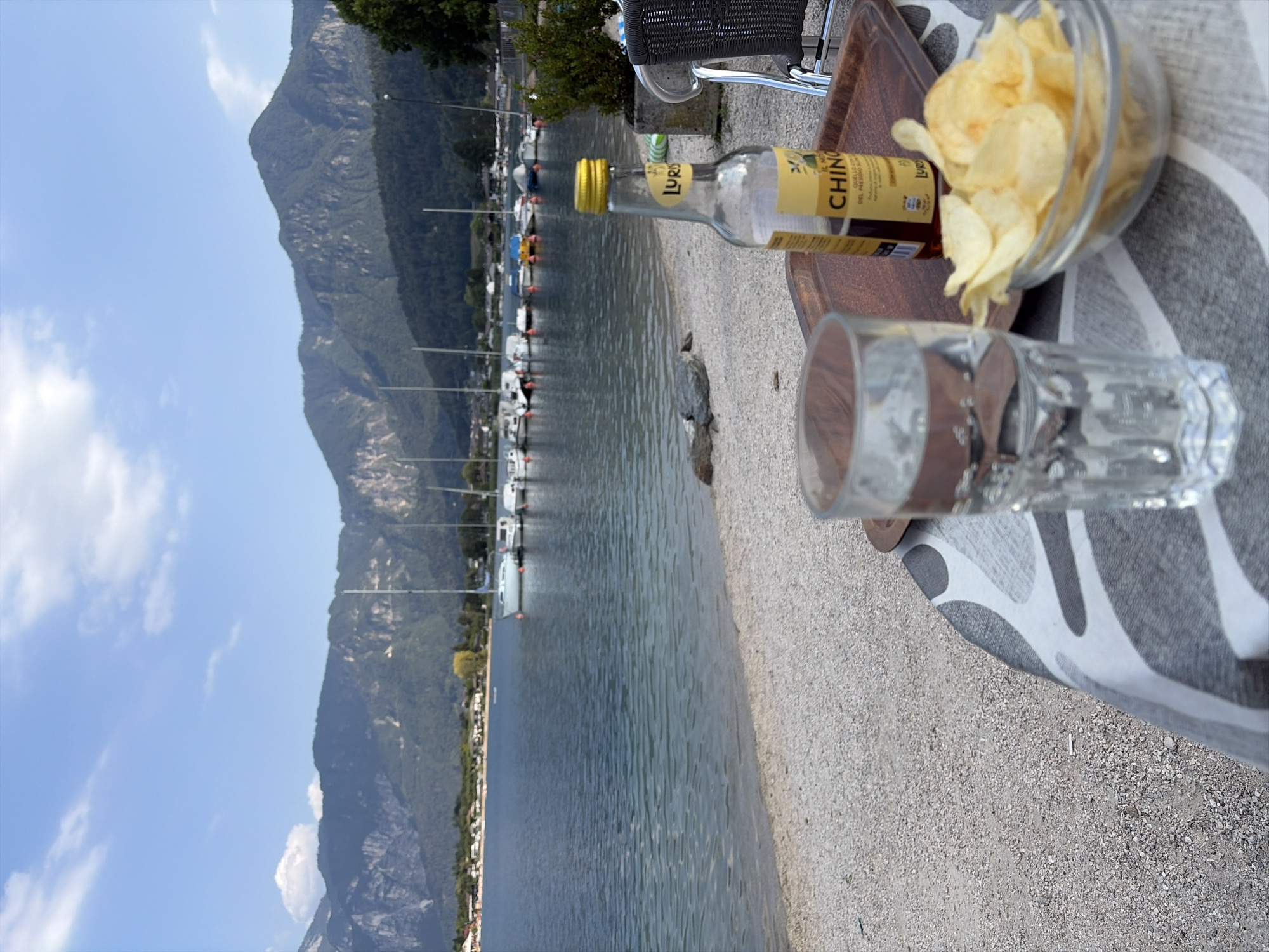This is a medium-shot, sideways photograph taken outdoors. The scene features a body of water, likely a lake, with a mountain range in the background. Several sailboats are moored on the water. In the foreground, on a gravelly surface, are a clear glass with water, a glass bottle with a yellow cap and label, and a clear glass bowl filled with potato chips. A wooden cutting board is underneath the bottle and bowl. A patterned fabric, possibly a tablecloth or blanket, is laid out on the gravel. The sky is blue with scattered white clouds. The lighting suggests it is daytime, with the sun casting shadows. The setting appears to be a lakeside leisure area. The label on the bottle is partially visible and reads "LURO" and "CHINO". There is no clear indication of people or a specific event taking place, suggesting a moment of relaxation.