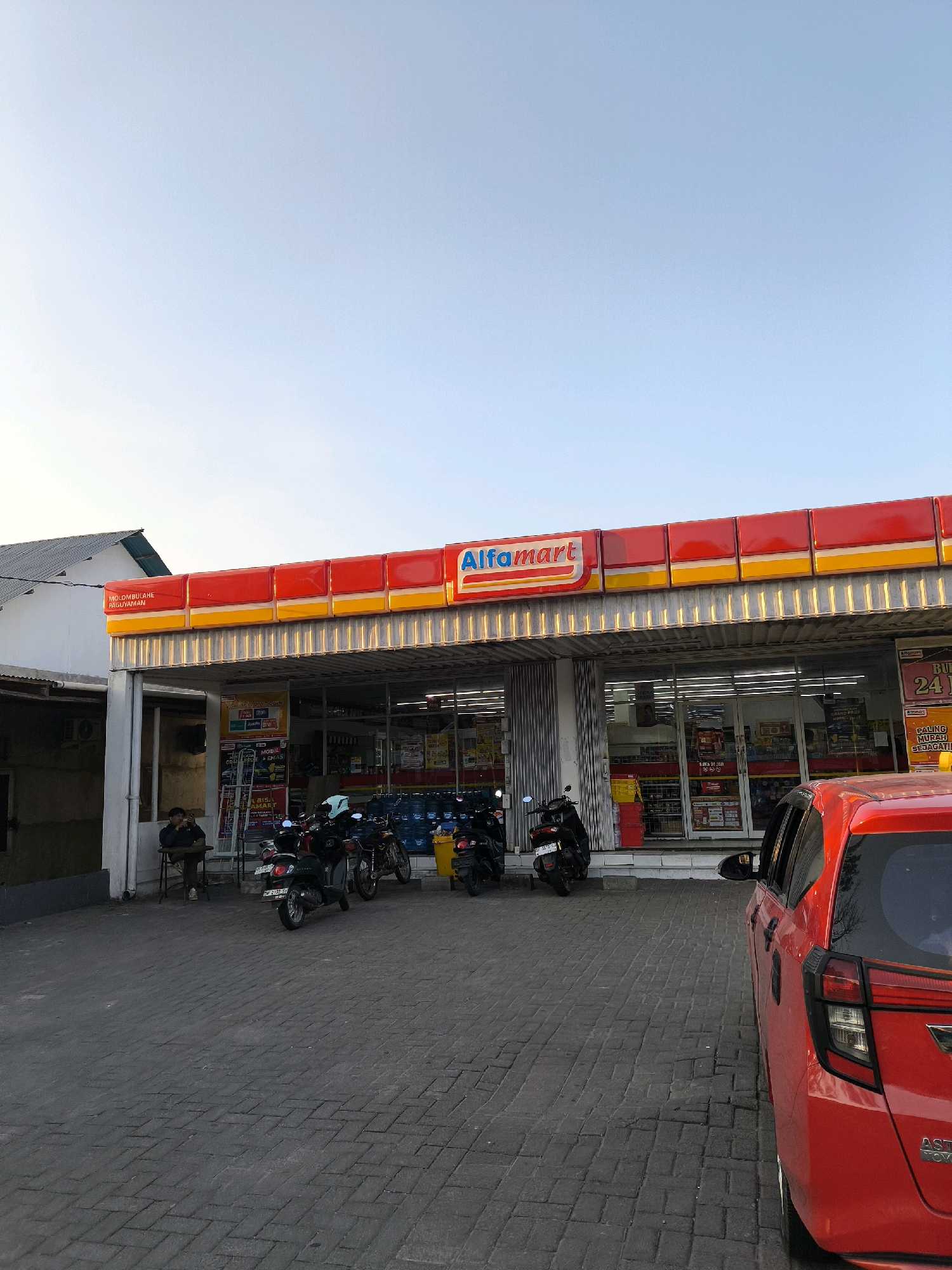 An exterior, eye-level, medium shot captures a bright red, four-door hatchback parked on a cobblestone lot in front of an Alfamart convenience store under a clear, pale blue sky in Tenilo, Indonesia. The store's awning, painted in red and yellow stripes, extends over the entrance, which features large glass doors and windows displaying shelves stocked with merchandise. Several motorcycles are parked in front of the store, and one person is seated at a table outside. The store has a prominent "Alfamart" sign. The overall impression is that of a typical Indonesian convenience store scene.