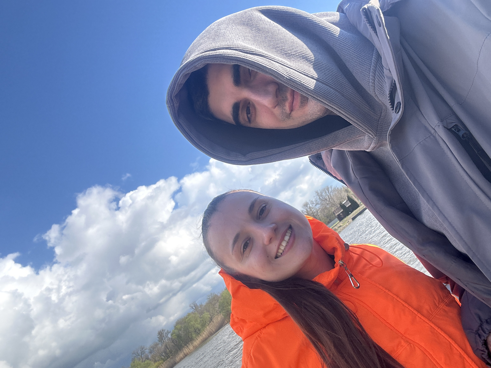 Two individuals are posing for a selfie outdoors. A man with dark hair is wearing a grey hooded jacket, the hood obscuring part of his face. A woman with long brown hair is wearing a bright orange jacket. Behind them, the background reveals a body of water, possibly a lake or river, with trees and vegetation along the shoreline. A partly cloudy blue sky is visible. The location is unspecified but mentioned as Ovoshchnoy, Russia.