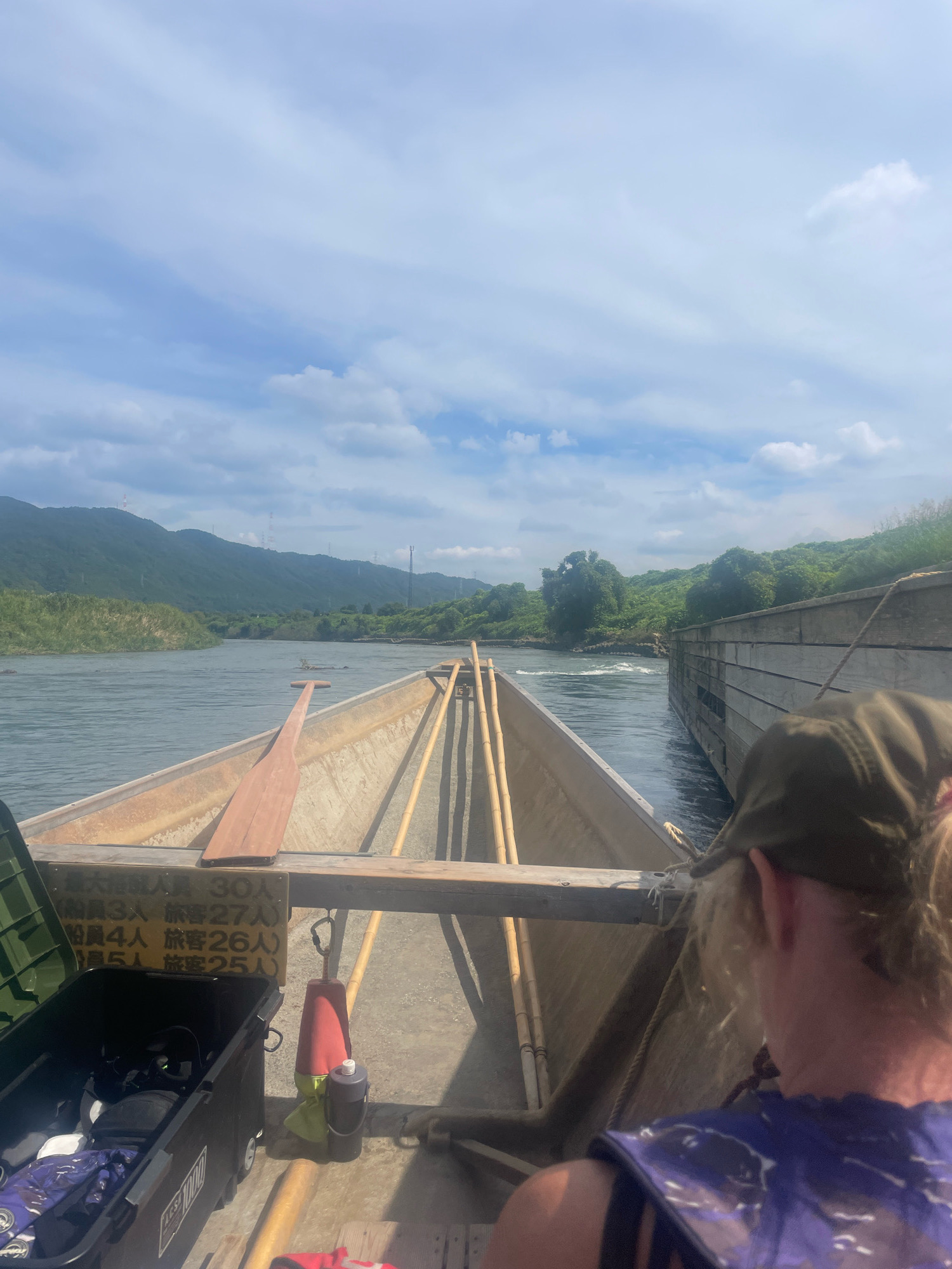 The image shows a view from the front of a boat on a river, likely in Kameoka, Japan. The boat appears to be a traditional wooden boat with two oars resting inside. A person wearing a green cap and a blue life vest is visible in the foreground, facing away from the camera. The river flows through a lush green valley, with mountains in the background. There is a wooden structure on the right side of the river, possibly a pier or a dam. The sky is partly cloudy. There is a sign inside the boat with Japanese writing that appears to indicate passenger capacity and age limits. A black storage container with items inside is also visible in the boat.