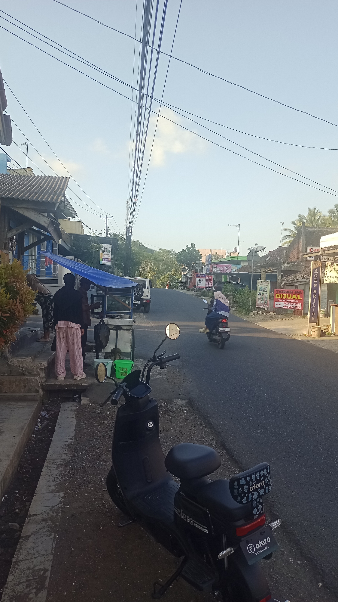 This image shows a street scene in Karyamandala, Indonesia, during daylight hours. In the foreground, a black electric scooter is parked on the side of the road. To the left, there is a sidewalk with some stairs leading up to a building. A cart covered with a blue tarp is present on the sidewalk, with two individuals standing nearby. One person is wearing striped pants and a dark top, while the other is wearing a dark jacket and pants. Another individual is partially visible in the background on the left.
The street itself is paved, and a few people on a motorcycle are riding away from the viewer. Buildings line the street, some with shop fronts and signage. A white SUV is parked on the side of the road further down. Power lines are visible strung across the sky above the street. The environment appears to be a residential or commercial area with a mix of shops and houses. The sky is clear with a hint of clouds.