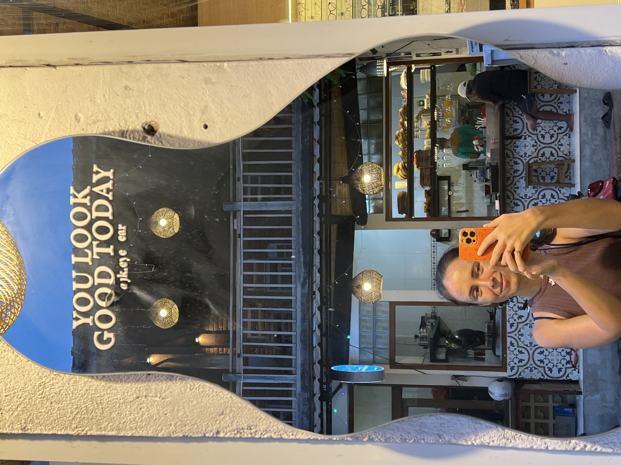 A person is taking a selfie in a uniquely shaped mirror with a white frame. The mirror reflects the exterior of a building with a sign that says "YOU LOOK GOOD TODAY" and "@jk.eye ear." Also visible in the mirror are wicker light fixtures, a building facade with a set of stairs, and a partial view of what appears to be a restaurant or bar setting with people inside. The person taking the selfie is holding an orange-colored smartphone. The reflection also captures a glimpse of patterned floor tiles and an interior space of a building. The location is likely in the Ubud District, Indonesia, based on the sign and architectural styles visible.