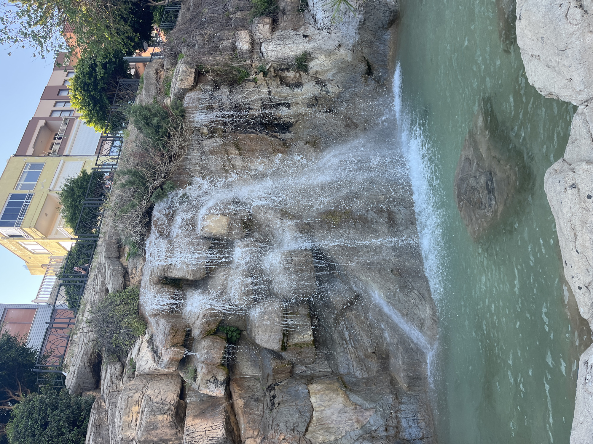 This image captures a vibrant scene centered around a multi-tiered waterfall cascading down a rugged, rocky cliff face. Water splashes and foams as it descends into a tranquil pool of light greenish-blue water at the base. Large, light-colored boulders frame the pool on the right.
The cliff itself is composed of various shades of grey and brown rock, with scattered patches of green foliage, dry bushes, and small trees clinging to its ledges and crevices. Above the cliff, an urban environment is visible, featuring multi-story residential buildings with balconies and windows, some with white roll-down shutters. The buildings display facades in shades of cream, yellow, and brown. Black metal railings with decorative patterns run along the edge of the cliff, separating the built environment from the natural drop. Lush green trees and shrubs are interspersed between the buildings and along the cliff edge.
The scene appears to be set during daytime under a clear, bright sky, suggesting sunny weather. The lighting indicates late morning or early afternoon. A person's head and shoulders are subtly visible behind the railing on the cliff edge in one of the cropped hints, looking towards the waterfall. There is no visible text in the image.