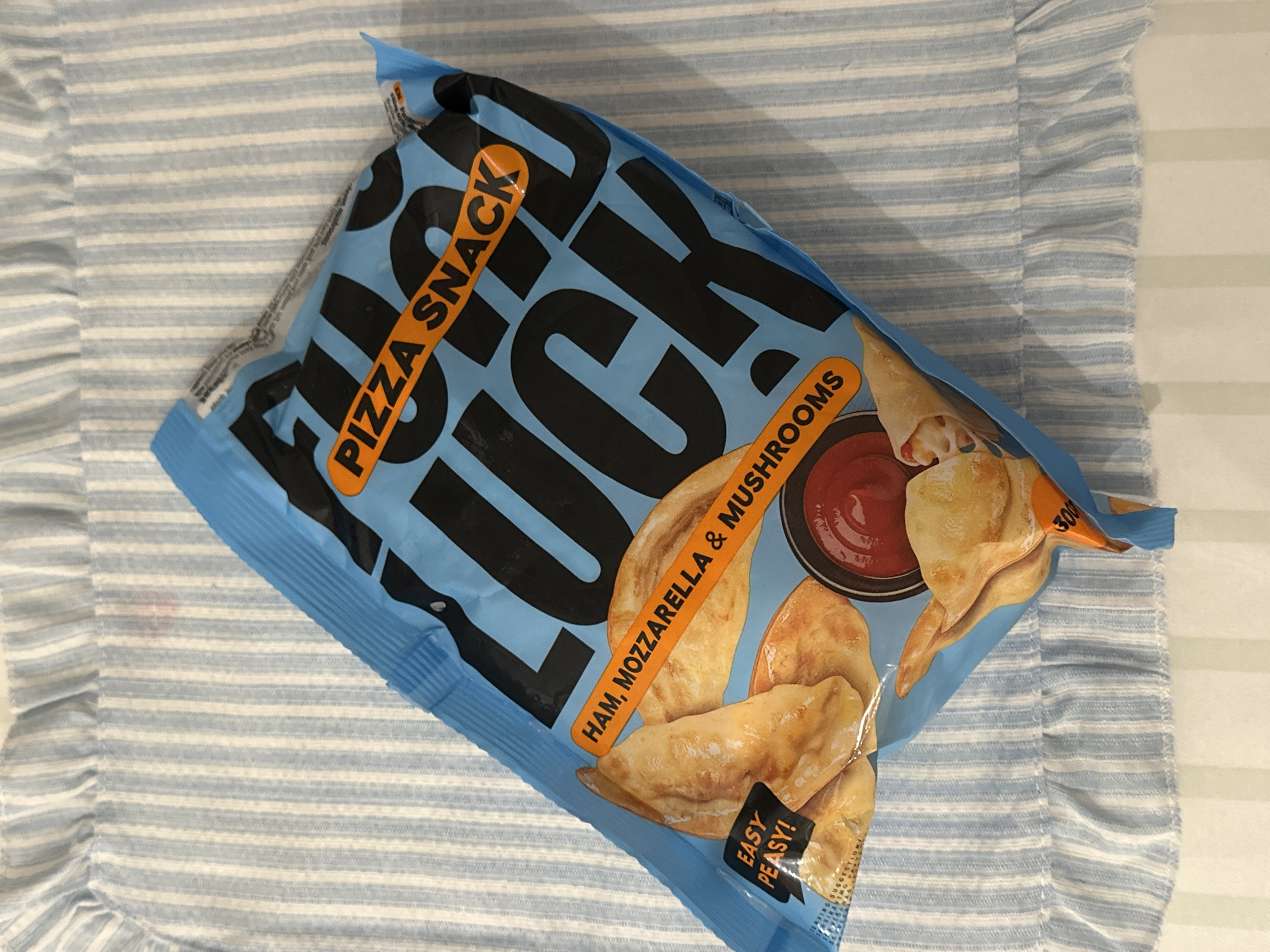 The image shows a bag of "Taste Luck" pizza snack with ham, mozzarella, and mushrooms. The bag is blue with black lettering and an orange accent. It is placed on a light blue and white striped fabric. There is no indication of a location in Bar, Montenegro, or any people present. The scene is a still life of a snack item.