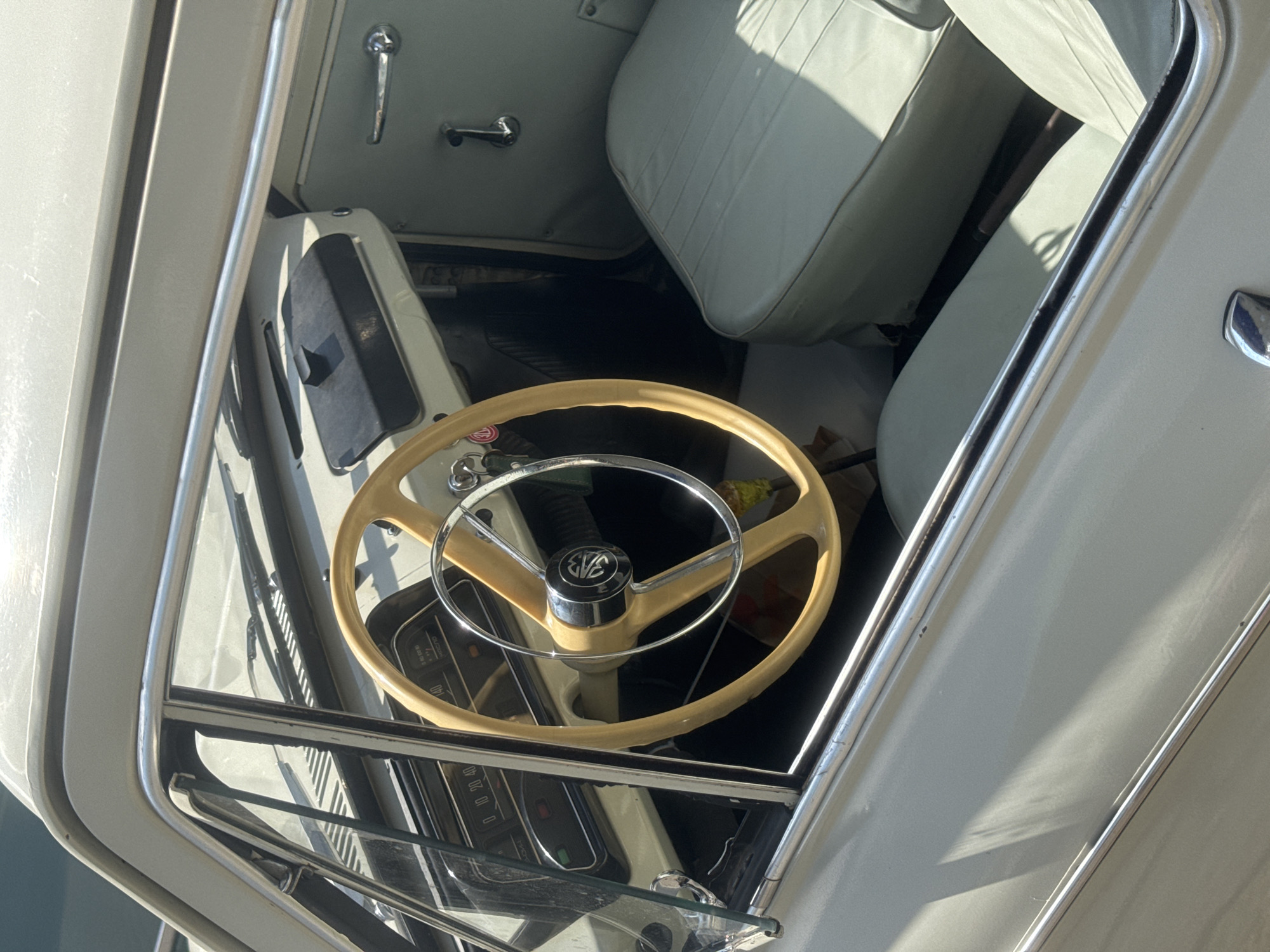 A close-up shot, taken from outside the open door, displays the interior of a vintage car. The beige steering wheel, with a chrome emblem, is prominently featured in the foreground. Behind the steering wheel, the car's dashboard and speedometer are partially visible. The car's seats are upholstered in light-colored vinyl. The car's interior and exterior are in good condition. The location is not discernible from the provided image.