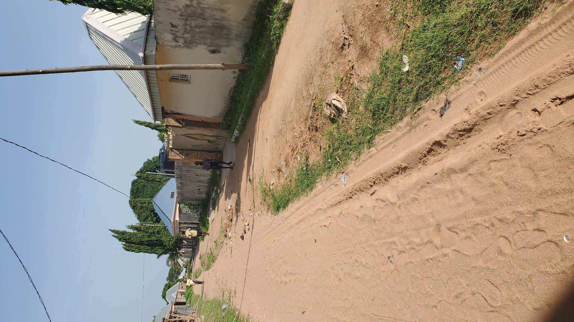 The image depicts a dirt road in Jalingo, Nigeria, with a few individuals walking along it. The road is unpaved and sandy, with tire tracks and footprints visible. On the right side of the road, there is a low, light-colored wall with a metal gate. Behind the wall, a building with a light-colored, sloping roof is visible. To the left of the road, there are some smaller structures and vegetation. The sky is clear and blue, suggesting it is daytime. One individual is walking towards the viewer on the road, wearing a checkered shirt and shorts. Further down the road, two more individuals, possibly children, are walking away from the viewer. The environment appears to be a residential area or a settlement on the outskirts of the city, characterized by its informal infrastructure and natural surroundings. There is some scattered debris and grass growing along the sides of the road.