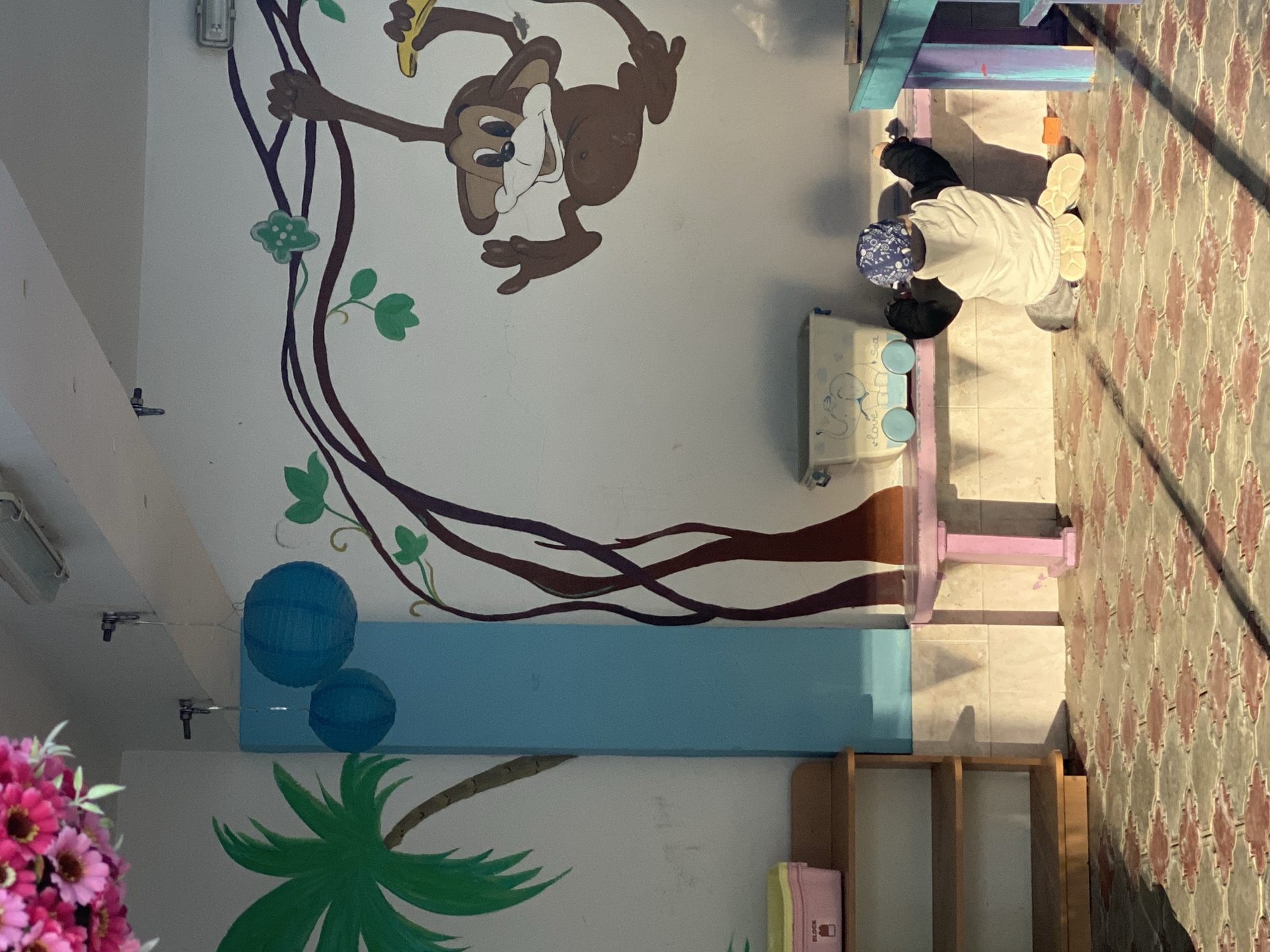 A brightly lit indoor room, likely a child's play area or classroom, features colorful murals and child-sized furniture. The main wall displays a large cartoon monkey, brown with a tan face, hanging from dark brown vines adorned with green leaves, holding a yellow banana. Towards the bottom left, a green palm tree frond is painted on the wall.
In the foreground to the right, a person, possibly a child, kneels on a reddish-brown and beige patterned tiled floor. They are dressed in a white long-sleeved shirt, black pants, and a blue and white patterned head covering. The person is engaged with a small, light-colored cart or toy box that has blue details and wheels. Faint text on the cart reads "i love sea," accompanied by a drawing, possibly an elephant. Strong shadows are cast across the tiled floor by the furniture, suggesting ample natural light, likely during the day.
Behind the kneeling person, a small table or bench with blue and purple painted elements is visible. A light blue wall section spans horizontally across the mid-ground, beneath the monkey mural. Above this section, two blue spherical paper lanterns hang from the ceiling. On the far left, a bouquet of vibrant pink flowers is partially visible, while a wooden shelf unit containing various items, including a pink box potentially labeled "BLOCK," stands further right. The scene conveys a playful and inviting atmosphere, characteristic of a child-centric environment.