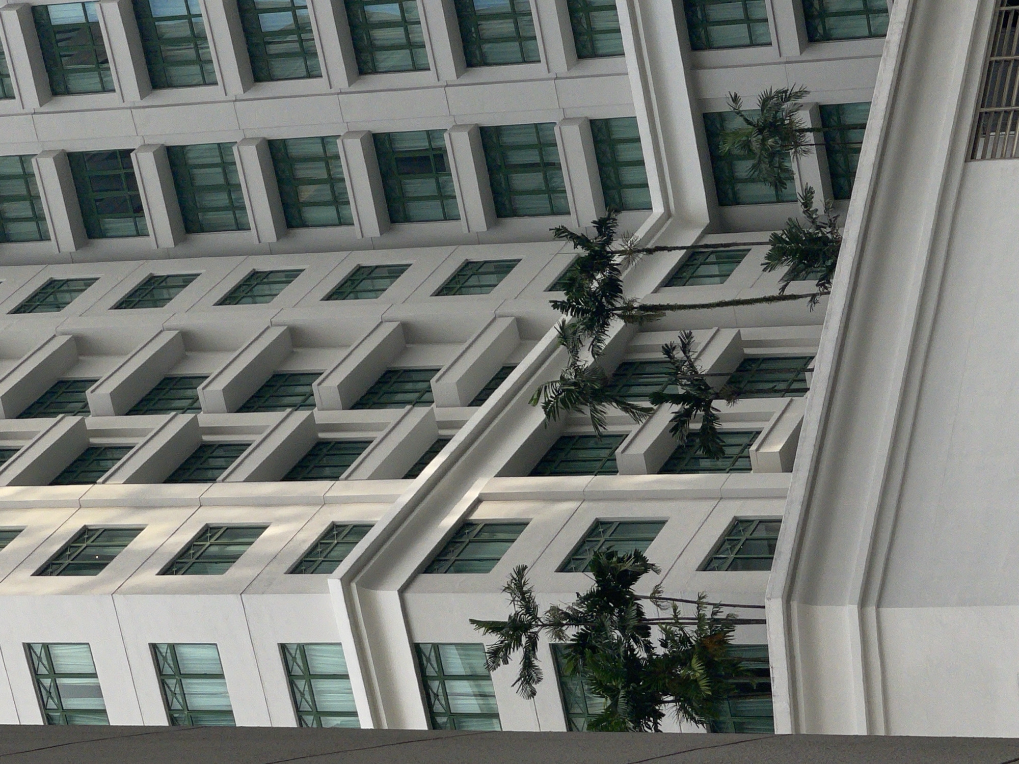 The image shows a close-up of a modern, white high-rise building in Kuala Lumpur, Malaysia. The building features repeating rows of windows with green frames, recessed within white overhangs. Several palm trees are visible growing from terraces or planters along the building's facade. The sunlight casts shadows on the building, highlighting its architectural details. The perspective is from a low angle looking upwards, emphasizing the height and scale of the structure. There are no people or vehicles visible in the frame. The overall impression is one of contemporary urban architecture.