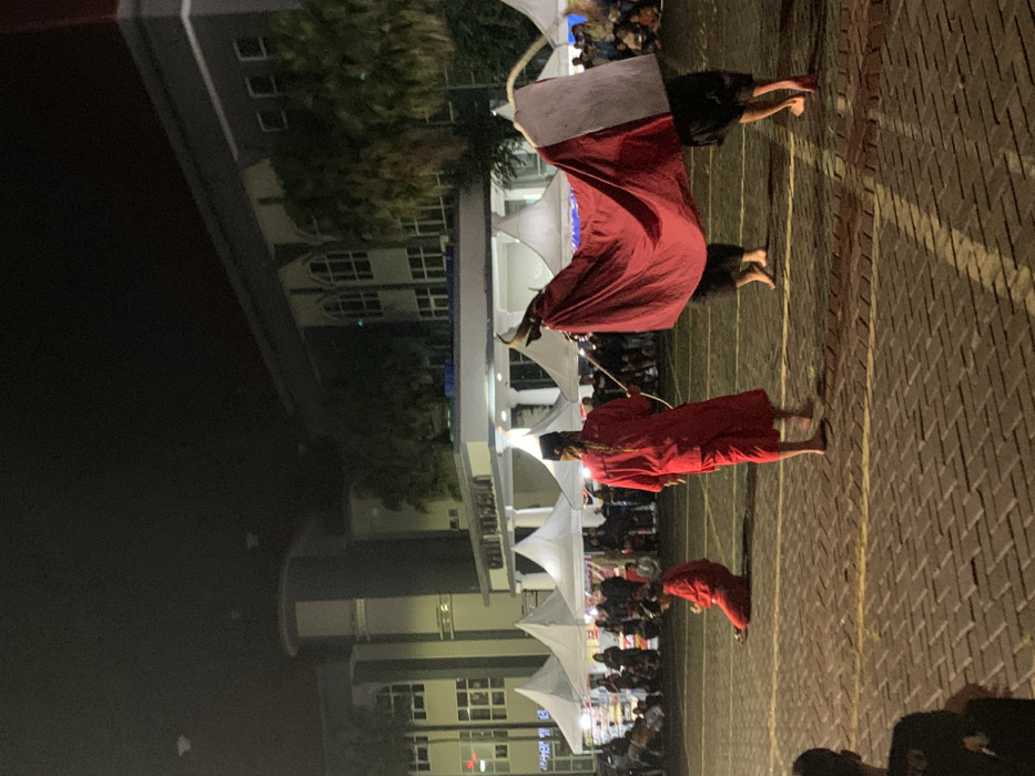 The image shows a performance in an outdoor space in Kecamatan Batu, Indonesia, at night. There are a group of people, some in red costumes and others in black, performing a traditional dance. In the foreground, two men are holding a large red cloth, possibly a costume, as a third man kneels on the ground. The performance is taking place in front of a large building with windows and a white awning. The building appears to be a government building. There are numerous white tents in the background, and the space is illuminated by streetlights. The ground is paved with red bricks.
