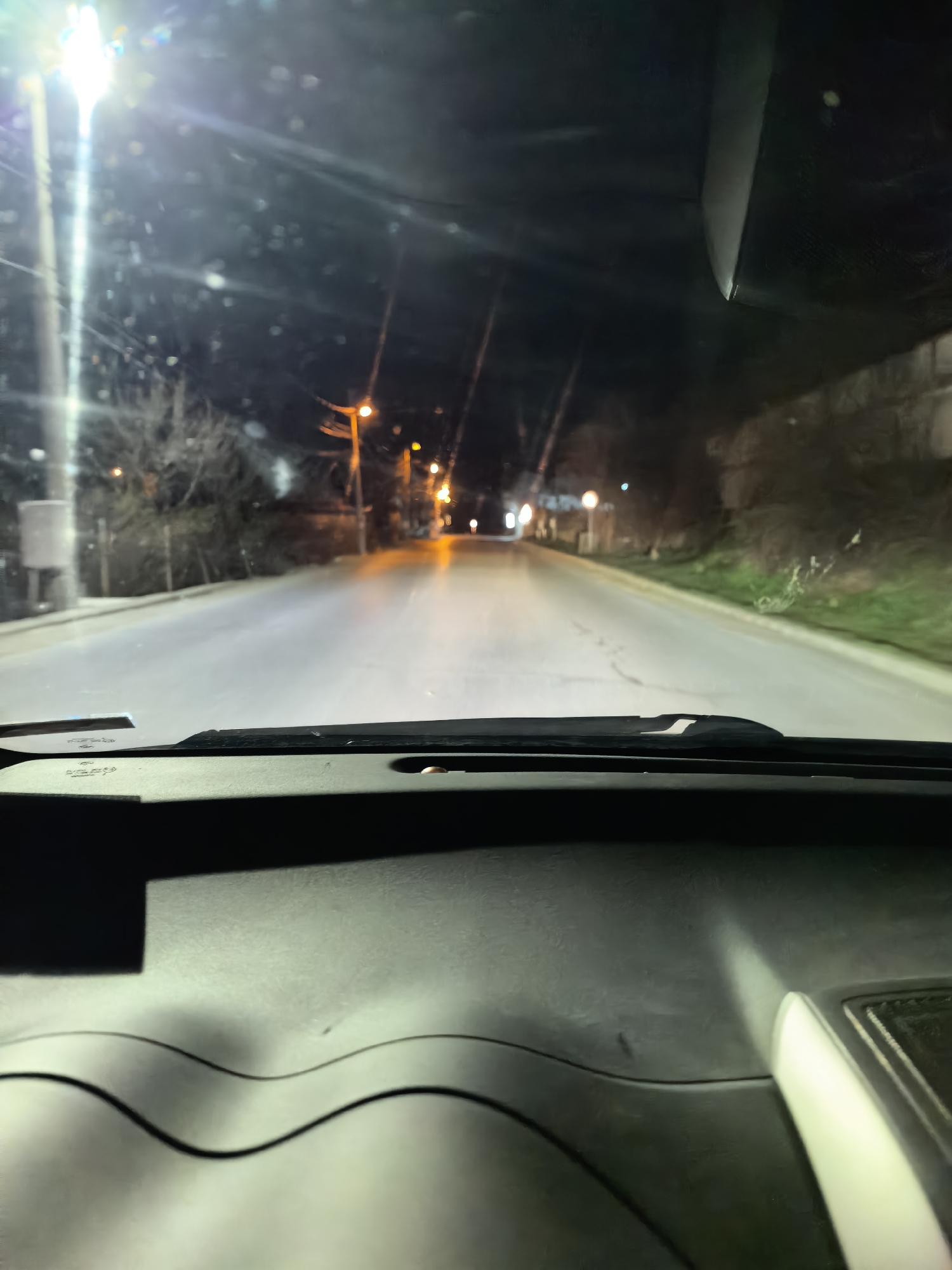 The image shows a nighttime street scene from inside a vehicle. The dashboard is visible in the foreground, with a road stretching into the distance. Streetlights illuminate the road, creating reflections on the wet pavement. Trees and buildings line the sides of the street. The location appears to be a residential or suburban area of Unknown, Unknown. No people are visible.
