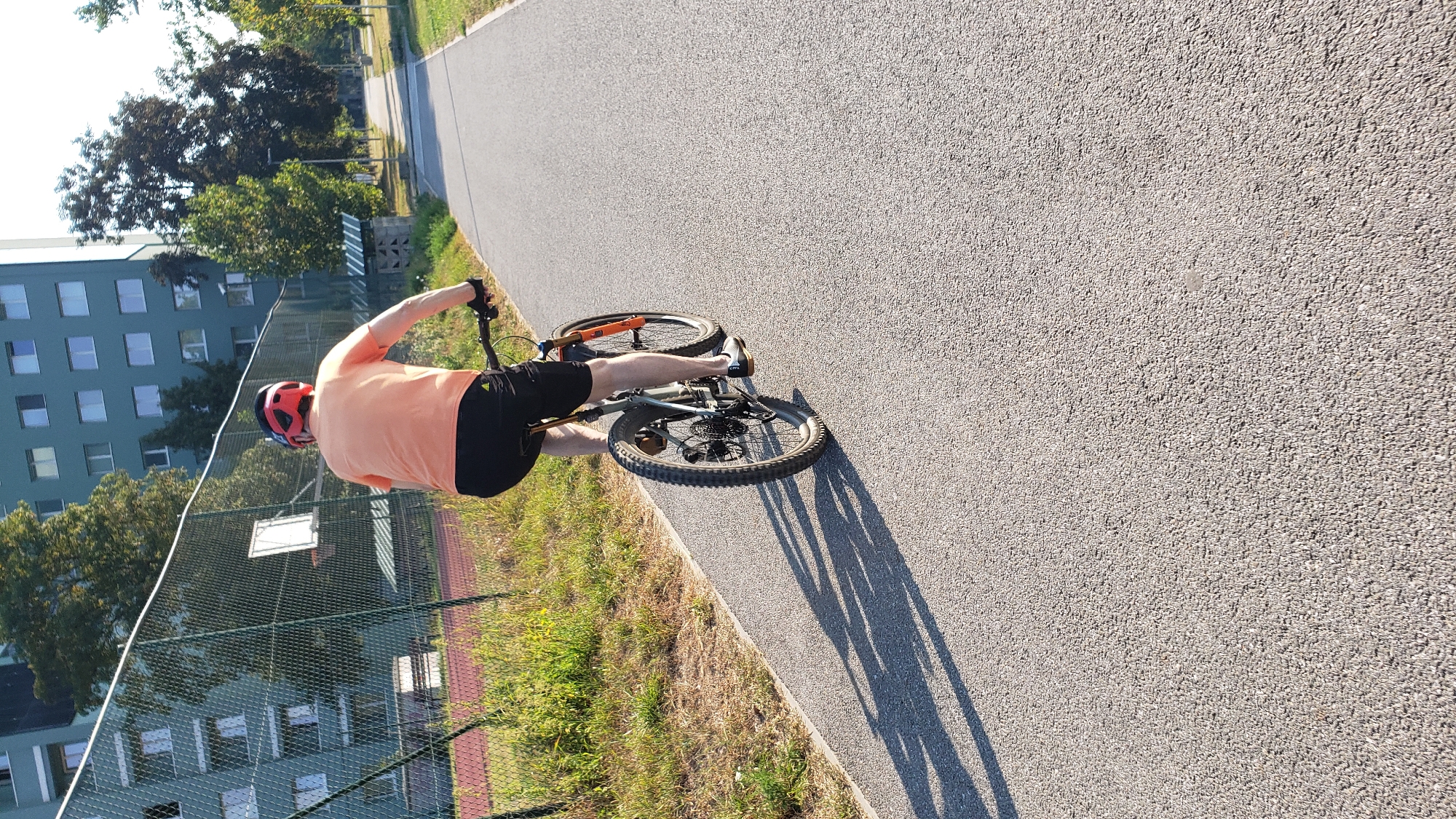 A person wearing a red helmet and an orange t-shirt is riding a bicycle on a paved path. The person is wearing black shorts and black cycling shoes. The bicycle is a mountain bike with knobby tires and orange accents on the front fork. The path is bordered by grass and a chain-link fence. Behind the fence, there is a green building with multiple windows. In the distance, there are trees and lampposts. The sun is casting a long shadow of the person and bicycle onto the path. The scene appears to be in a park or recreational area within the city of Prešov, Slovakia.