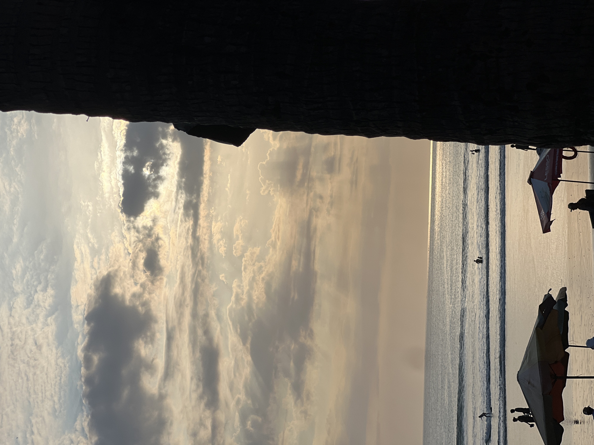This is a vertical, medium shot of a sunset at Kuta Beach, Indonesia. In the foreground, the dark silhouette of a palm tree trunk occupies the right side of the frame. To the left, two beach umbrellas are visible, one in the foreground with a muted color palette, and another partially visible in the mid-ground. Beyond the beach, the ocean stretches to the horizon, with gentle waves rolling in. Several figures are dispersed across the sand and in the water, some appearing to be surfing. The sky is filled with a mix of clouds, with sunlight breaking through, creating rays of light that illuminate the scene. The overall atmosphere is serene and warm, typical of a tropical sunset.