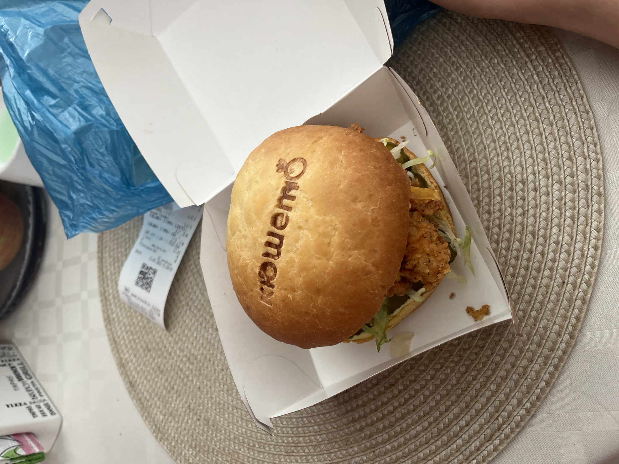 A close-up, high-angle shot shows a chicken sandwich in an open, white cardboard box. The top bun of the sandwich has the word "KFC" imprinted on it. Lettuce and fried chicken are visible within the sandwich. The box sits on a round, woven placemat. A partially visible receipt with a QR code is to the left of the box. Part of a blue plastic bag is visible in the upper left of the frame. The location appears to be somewhere in the city of Blagoevgrad, Bulgaria, judging from the Cyrillic lettering on the burger.