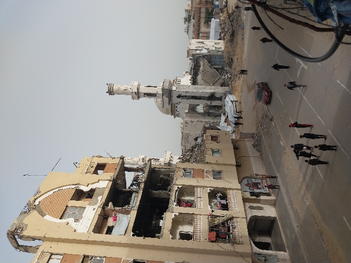 This image depicts a street scene in Khan Yunis, Palestinian Territory, characterized by extensive structural damage. In the foreground on the left, a multi-story, light-colored residential or commercial building shows significant destruction, with large sections of its facade missing, exposing burnt and rubble-filled interiors on upper floors. Balconies on the lower floors still have clothes hanging to dry. Directly across the street, in the mid-ground, a damaged mosque is visible, with its white minaret largely intact but the main prayer hall and dome extensively collapsed into debris.
The street itself is asphalt with some visible white markings, appearing mostly clear but bordered by rubble and dirt. A group of approximately ten young males is gathered in the center-left of the street, some interacting while others stand nearby. A single red sedan is parked or driving slowly on the right side of the street, heading away from the viewer. Several other pedestrians are scattered across the scene, walking individually. The background reveals more heavily damaged buildings and piles of debris, under a uniform, pale gray sky. A dark, rusted metal structure is partially visible in the extreme lower right foreground.