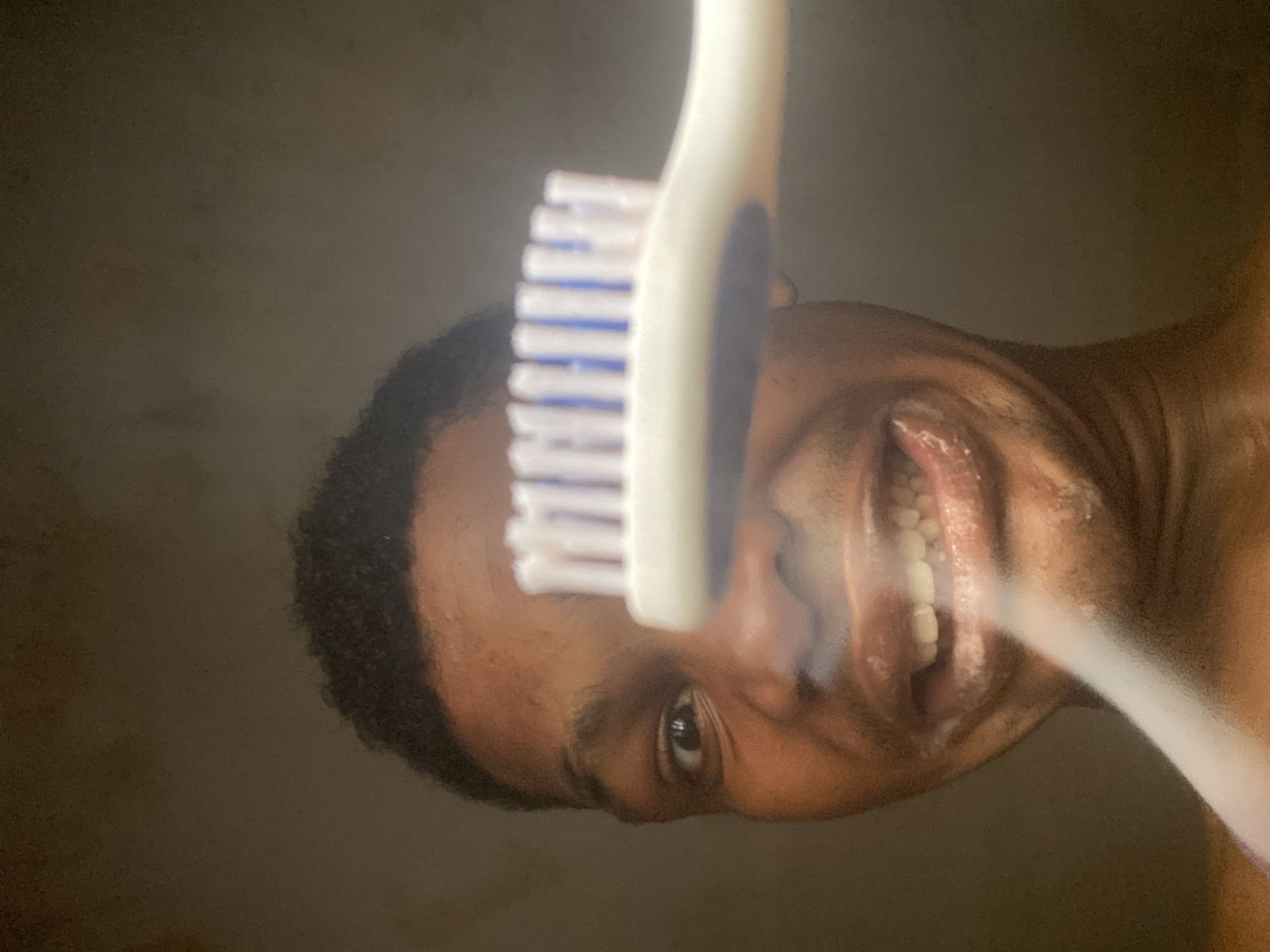 A man is holding a toothbrush with white and blue bristles close to his face. His expression is a wide smile revealing his teeth. He is an African man with dark hair, wearing a brown shirt. He is located in Ado, Nigeria. The background is a dark, neutral color.