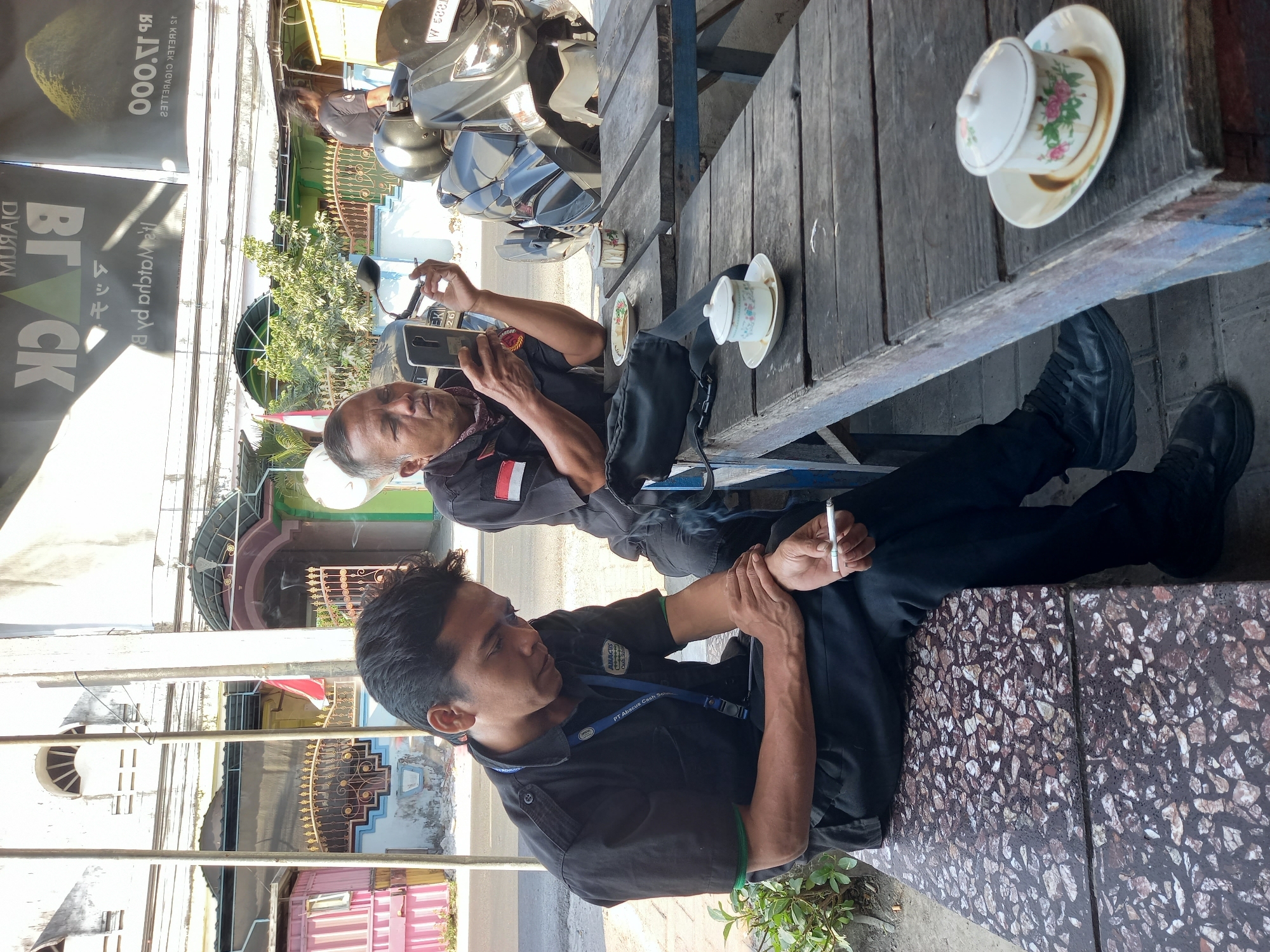 The image depicts a daytime outdoor scene in the city of Bungah, Indonesia. Two men are present, seated at wooden tables. The man on the left, wearing a black uniform shirt with an "ABACUS Cash Services" logo and a blue lanyard, is looking down and holding a lit cigarette. The man on the right, also in a black shirt, is holding a smartphone in his left hand and a lit cigarette in his right hand, appearing to be smoking. A motorcycle is parked behind the man on the right. On the tables are two small cups with saucers, one containing a dark liquid. The environment shows a street in the background with buildings and foliage.