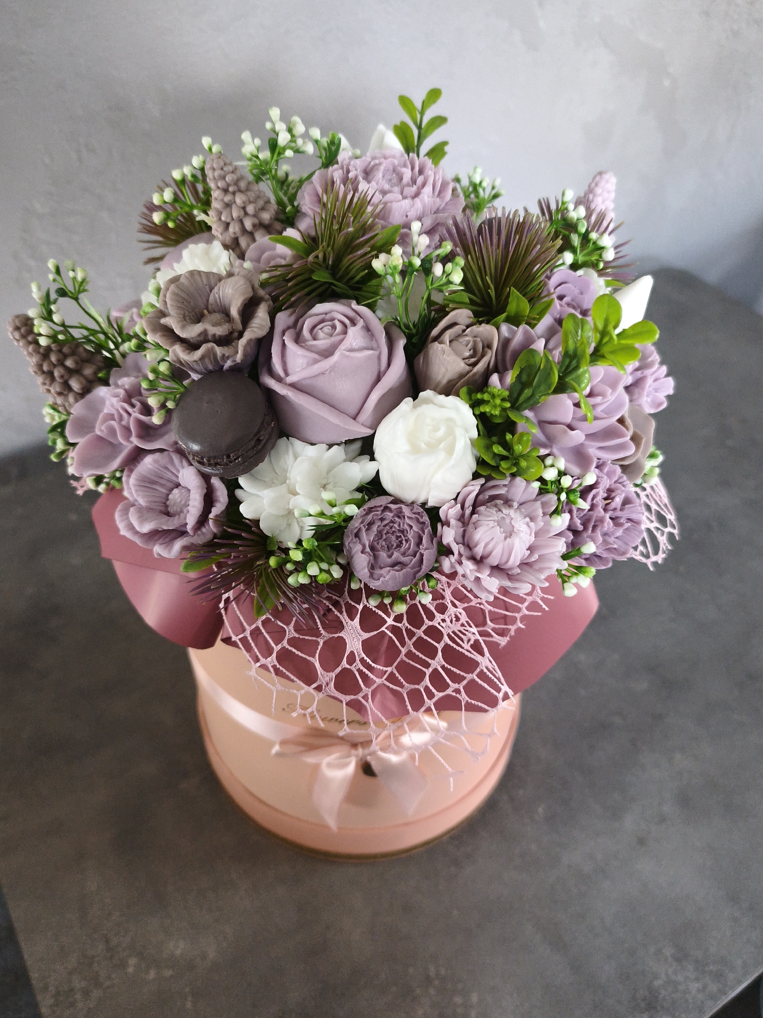 This media file features a close-up, overhead shot of a decorative arrangement of soap flowers presented in a round, pink gift box. The arrangement is a mix of various floral shapes and sizes, predominantly in shades of lavender, mauve, and cream, with some brown and white accents. Amongst the flowers are small green leaves and sprigs, as well as what appears to be a dark brown macaron. The gift box itself is a soft pink with a satin ribbon tied in a bow at the front, and it is partially wrapped in a delicate, light pink mesh netting. The background is a textured, grey wall. The lighting suggests an indoor setting, possibly a studio or home, with no indication of time of day or weather. There is no visible text in the image. The overall impression is one of a handcrafted, artistic gift.