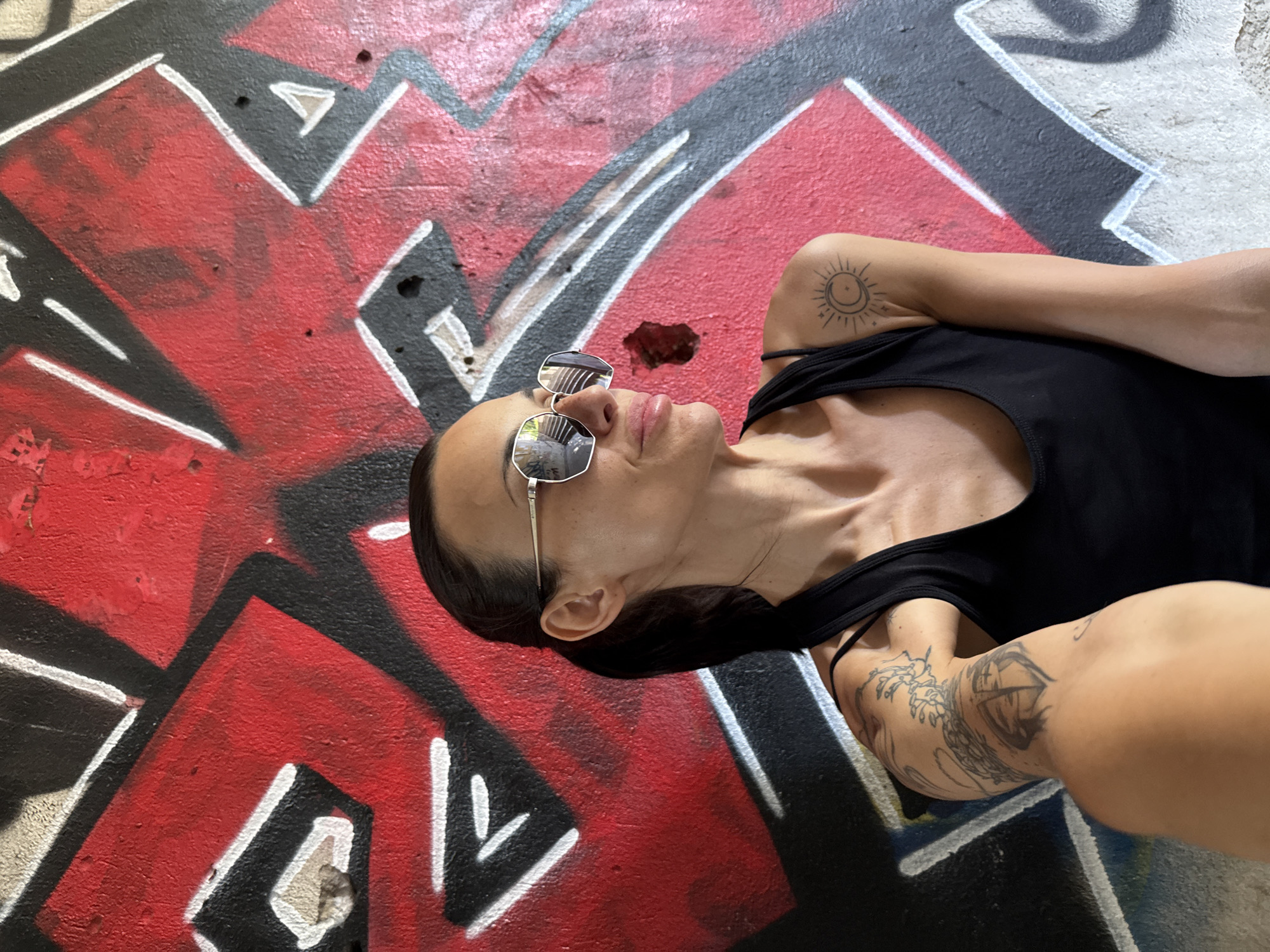 The image shows a woman with dark hair pulled back in a ponytail, wearing sunglasses and a black tank top, standing in front of a graffiti-covered wall. She has tattoos on her left arm, including a dragon and a shield, and a sun and moon tattoo on her right shoulder. The graffiti on the wall is primarily in red and black, with white accents. The woman is looking to her right and appears to be taking a selfie. The background suggests an urban or industrial setting, possibly an underpass or abandoned building. The photograph was taken in Pleven, Bulgaria.