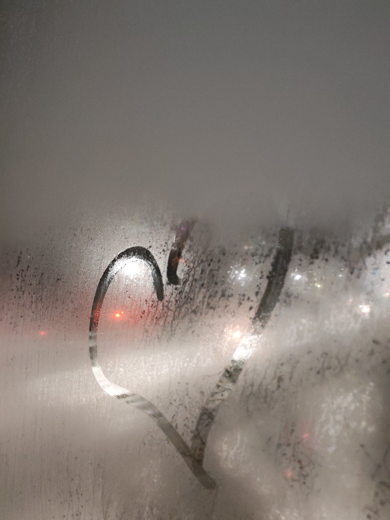 A heart shape is drawn into the condensation on a glass surface. Outside the glass, light sources are visible in a blurred fashion, indicating an external environment such as nighttime. The condensation on the glass is uneven, with droplets and streaks obscuring the view. The lack of sharpness and detail suggests the photo was taken from inside a building. This image contains no evidence to determine it's specific location in Izhevsk, Russia.