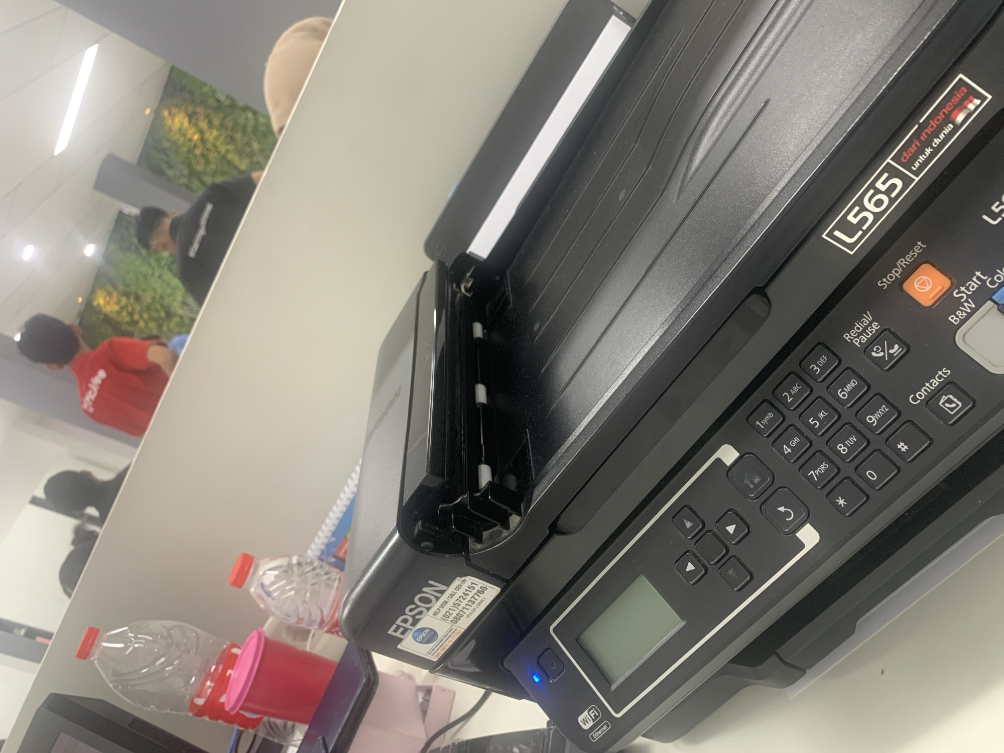 The image shows a close-up view of a black Epson L565 printer on a white desk. The printer has a control panel with a small LCD screen and several buttons, including a numerical keypad and navigation keys. A blue Wi-Fi indicator light is visible on the left side of the printer. An Epson service sticker with contact information is attached to the top left of the printer. In the background, slightly out of focus, are several water bottles, one red container, and a blurred reflection of people in what appears to be an office environment. One person in the reflection is wearing a red shirt with white lettering, and another is wearing a black shirt. The overall setting suggests an indoor workspace, possibly an office in Palembang, Indonesia, given the presence of text in Indonesian on the printer's sticker and a small flag graphic next to "dari indonesia untuk dunia".