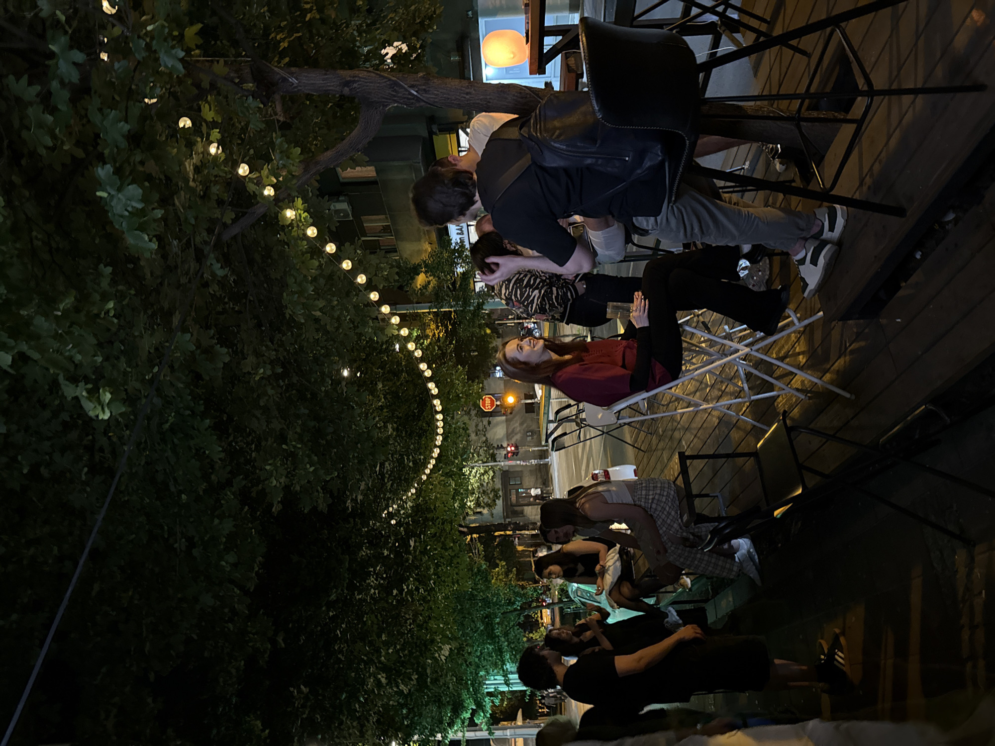 The image depicts an outdoor scene in Belgrade, Serbia, at night. A string of lights illuminates a tree on the left, leading toward a street with a stop sign and traffic lights. Several people are gathered around tables and chairs on a wooden deck, suggesting an outdoor bar or restaurant setting. Some individuals are standing, while others are seated, and they appear to be conversing. One person, with a black shirt and a black backpack, seems to be talking to someone sitting on a white chair. The overall atmosphere is relaxed and social, with the city visible in the background.