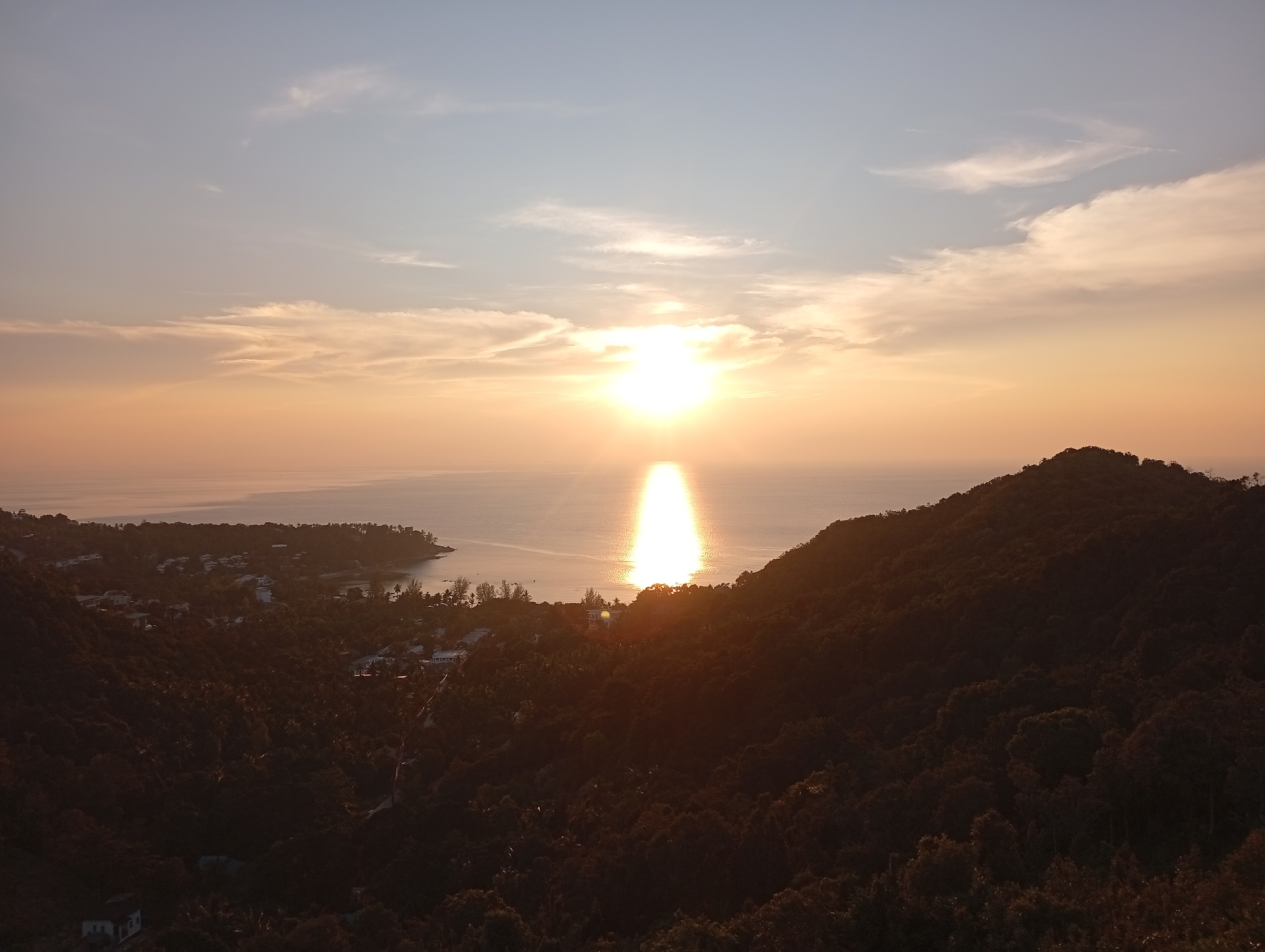 The media file presents a panoramic view of a tropical coastal landscape at sunset, likely captured from a high vantage point overlooking Baan Wang Ta Kien, Thailand.
The central and most striking feature is the sun, a brilliant, glowing orb positioned low on the horizon over the expansive ocean. It casts a vivid, shimmering column of light directly across the water surface towards the viewer, creating a dramatic reflection path. The sky above is painted with warm hues of golden orange near the horizon, gradually transitioning into softer tones of light blue and grey further up. Scattered, wispy clouds are visible, particularly in the upper regions, catching the ambient light.
In the foreground and midground, verdant hills covered in dense, dark green foliage dominate the lower portion of the image. These hills appear largely in silhouette due to the backlighting from the setting sun, enhancing the contrast with the bright sky and ocean. Along the winding coastline, which features several bays and headlands, scattered residential buildings or resort structures can be seen nestled among the trees.
No people are visible, and no specific activities are taking place other than the natural event of the sunset. The image conveys a serene and majestic atmosphere, highlighting the natural beauty of the landscape during golden hour. There is no visible text within the frame.