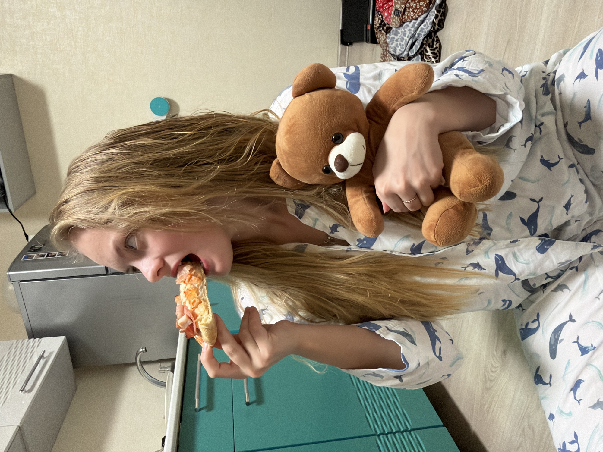 A young Caucasian woman with long blonde hair is eating a sandwich while holding a brown teddy bear. She is wearing pajamas with a whale pattern. She appears to be in a kitchen, as a sink, cabinets, and appliances are visible in the background. The location is Izhevsk, Russia.