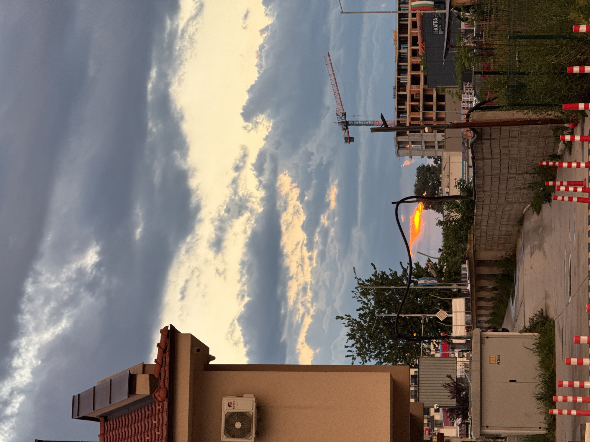 The image depicts a view of Plovdiv, Bulgaria, at dusk. In the foreground, a low building with a terracotta tiled roof and a beige wall with an air conditioning unit is visible. A street with road markings and red and white striped bollards runs along the right side. Beyond the building, a street scene includes foliage, utility boxes, and street signs. In the midground, a construction site features an unfinished building and a red crane. The sky is filled with dramatic clouds tinged with orange from the setting sun.