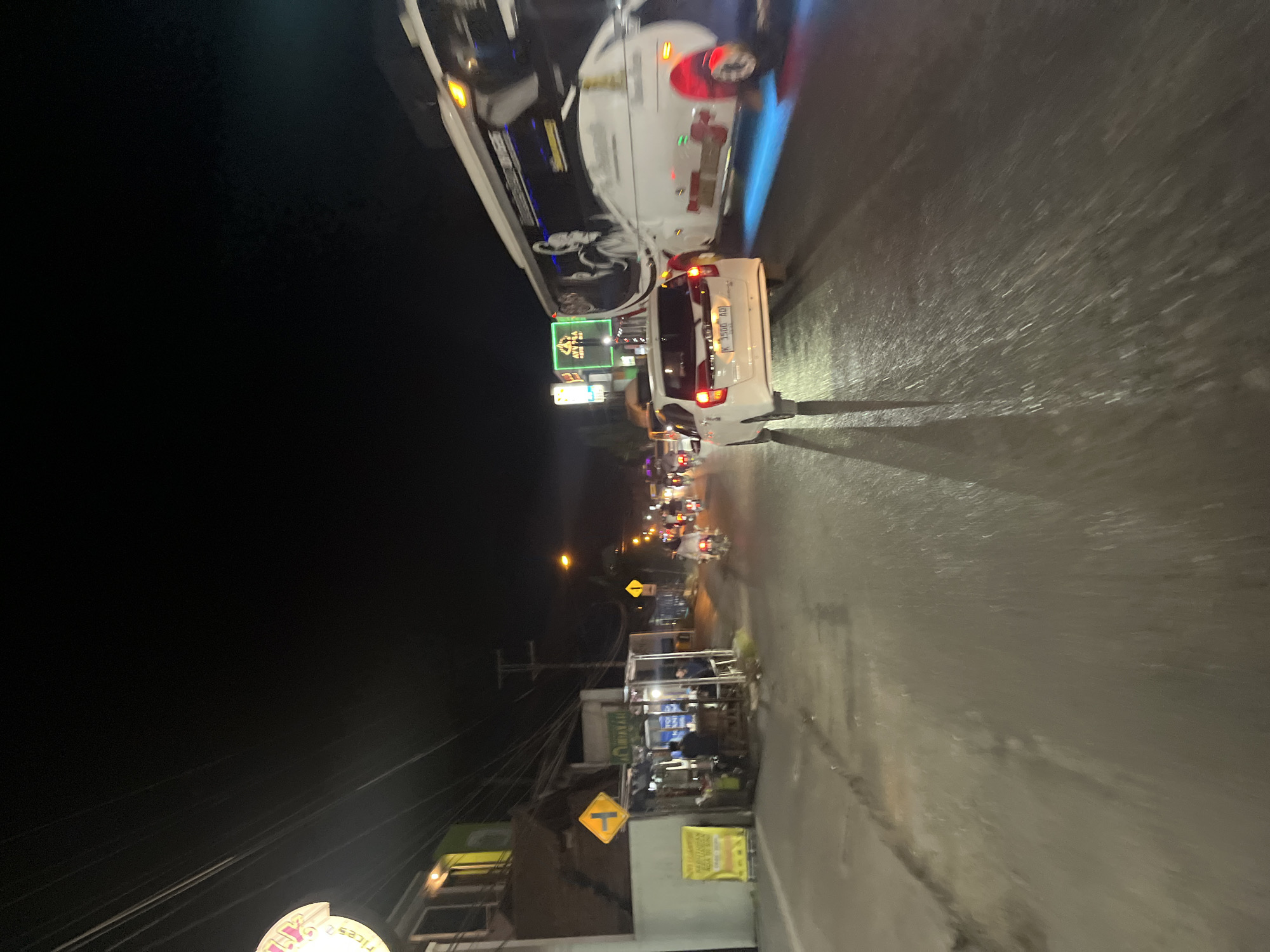 This is a nighttime, slightly blurry, eye-level photograph of a street scene in Banyuputih, Indonesia. The road is wet, reflecting the light from vehicles and street lamps. On the left side of the street, there is a low-rise building with a light-colored facade and windows. A yellow sign with a black arrow indicating a turn is visible. In front of the building, there are some stalls with people present. Further down the street, there are more stalls and streetlights.
In the center of the frame, a white car is driving on the road, its license plate reading "K 1500 ADI". To the right of the car, a large white bus is traveling in the same direction. The bus has graphics on its side and blue undercarriage lights. In the background, there are other vehicles, including motorcycles, and neon signs. The sky is dark, indicating it is nighttime. The overall atmosphere of the image is that of a busy street at night.