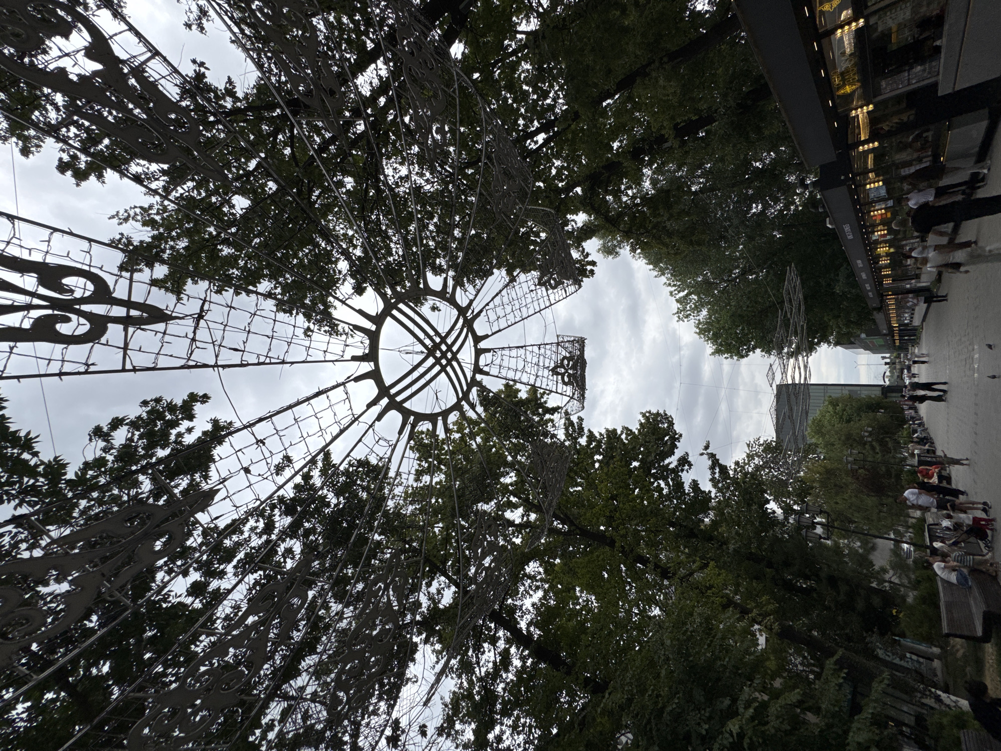 The image depicts a public promenade area in Shymkent, Kazakhstan, likely in a park or pedestrian zone. Overhead, a large, intricate metal structure resembling a sunburst or a stylized canopy is visible, interwoven with trees and string lights. The structure is ornamental, featuring decorative metallic elements against a cloudy sky.
Below the canopy, a paved walkway stretches into the distance, lined with benches and mature trees. On the left side of the walkway, there are wooden benches with people seated and standing, including children. On the right side of the walkway, a row of commercial kiosks or shops with illuminated displays is present, suggesting a commercial or entertainment area.
Several individuals are walking along the promenade. A group of people, including adults and children, are gathered near the benches on the left. Further down the promenade, more people are seen walking in both directions. The atmosphere appears casual and public. The overall environment is a blend of natural elements (trees, sky) and urban infrastructure (walkway, structures, kiosks). The time of day appears to be overcast, with diffused natural light.