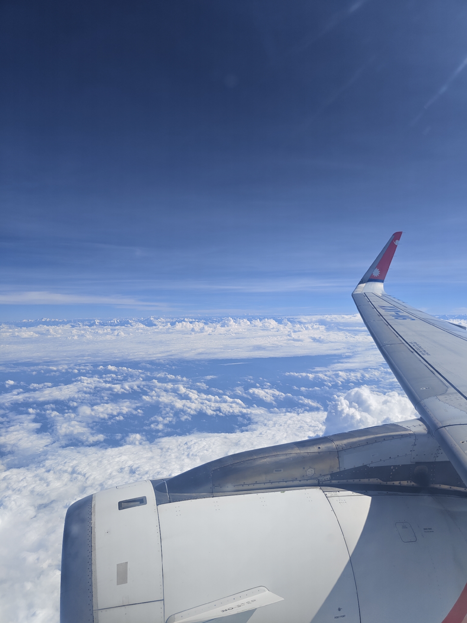 This is a photograph taken from an airplane window, looking out over a vast expanse of clouds. The sky above is a deep, clear blue. In the foreground, parts of the airplane are visible, including the engine cowling, which is white and metallic, and the right wing, which has a red tip with a national emblem. The clouds form a dense, white blanket beneath the aircraft, with the faint outlines of mountains visible in the distance, suggesting a high altitude flight. There are no people or specific city structures visible in this image, and therefore no discernible location in Shaktigarh, India.