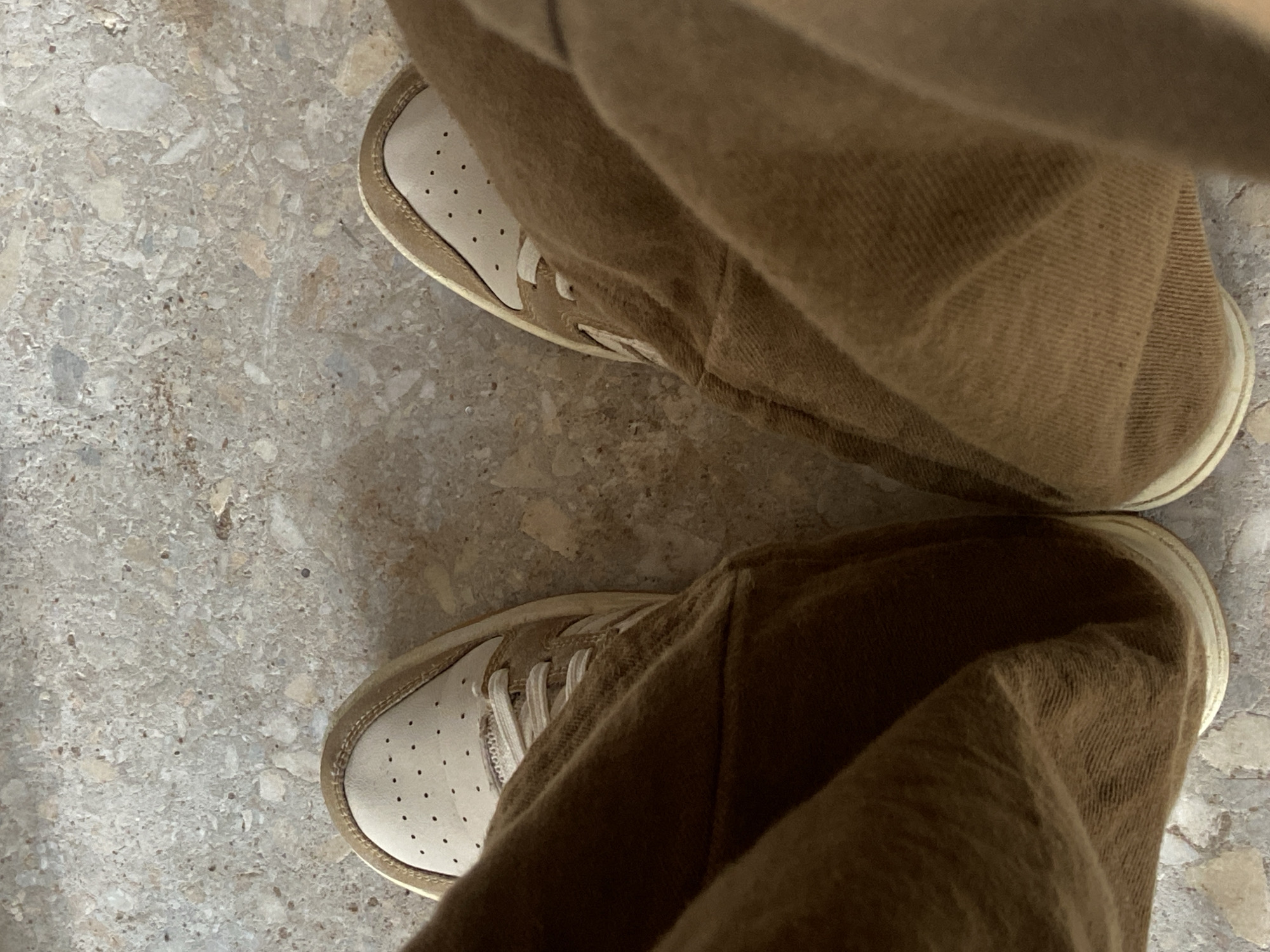 The image displays a top-down view of a person's lower legs and feet. The individual is wearing loose-fitting, medium brown trousers, made of a woven fabric with visible texture and creases, which rest over the tops of their shoes. On their feet are light-colored sneakers with an off-white toe box featuring multiple small, dark perforations, and a slightly darker tan or beige trim around the edges and lacing system. The shoes are fastened with off-white laces. The person is standing on a speckled floor surface, which appears to be terrazzo or a similar composite material, characterized by irregular, light-toned aggregate (in shades of white, gray, beige, and light brown) embedded within a lighter matrix. The lighting is even, suggesting an indoor or shaded outdoor setting. There is no visual information within the image to identify a specific geographic location, such as the city of Mararaba, Nigeria.