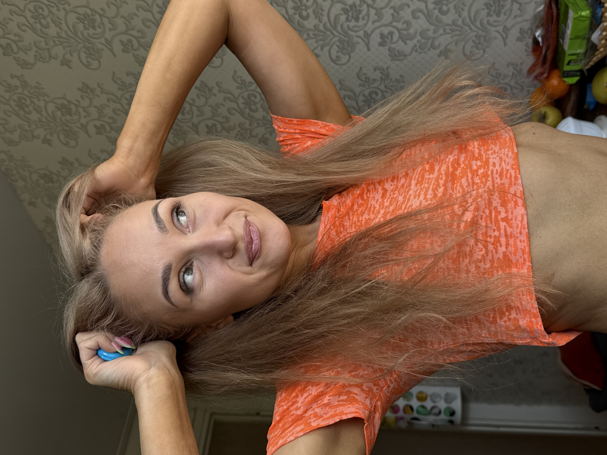 A young adult woman is captured in a close-up portrait, appearing to be lying down and looking upwards towards the camera with a subtle smile or smirk. She has long, straight, light blonde or light brown hair that flows around her head. Her eyes are light-colored, complemented by defined eyebrows and a touch of makeup. She wears a short-sleeved, orange cropped t-shirt with a light-colored, abstract pattern, revealing her midriff and highlighting her toned physique. Her arms are raised, with hands positioned near her head, framing her face. In her left hand, she holds a small, rounded blue object, and her fingernails are adorned with a pink and white manicure on some digits.
The background behind her features a wall covered in a light grey or beige wallpaper with an intricate, ornate floral or damask pattern. To her right side, the background is somewhat cluttered, showing various household items. These include visible green apples and oranges, a green package (possibly food), and a white tissue box or similar container. Further back, a small, white organizer box with multi-colored circular compartments is discernible. The setting suggests an indoor, casual home environment in Chaikovsky, Russia. The woman's pose and expression convey a confident and playful mood.