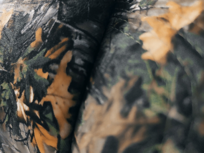 A close-up shot displays a camouflage pattern, predominantly featuring muted greens, browns, and hints of yellow, mimicking the appearance of foliage. The texture appears to be a fabric with intricate details. The colors blend, creating an organic, naturalistic visual effect. The image is slightly blurred, suggesting it might be a macro shot focusing on the texture rather than precise details.