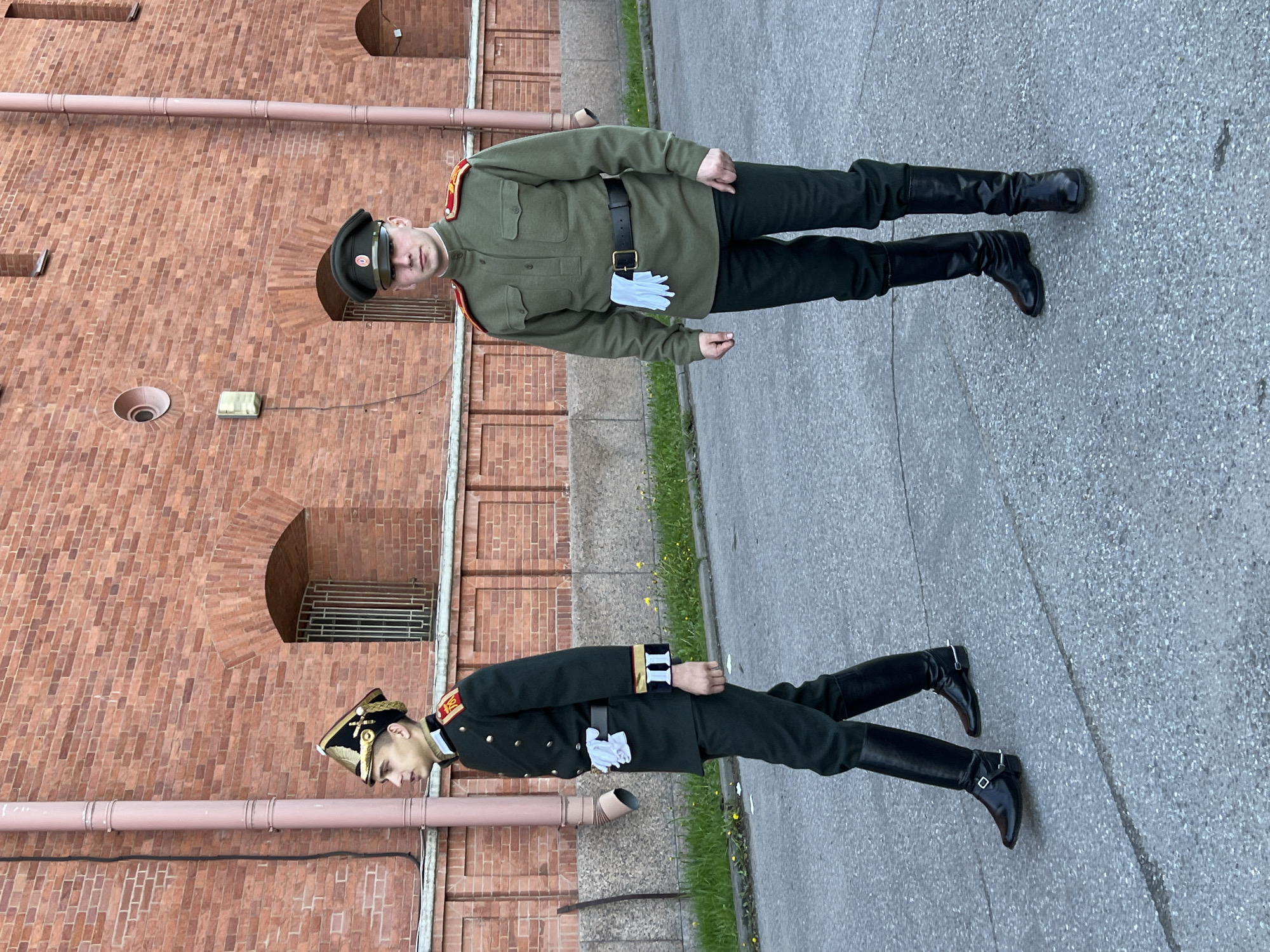 The image depicts two young men standing at attention in front of a red brick building. Both individuals are dressed in military uniforms, including black boots. The man positioned higher in the frame is wearing a light green uniform with red epaulettes. He is also wearing a black belt with a silver buckle and holding white gloves. The other man wears a darker green uniform with elaborate shoulder boards and white gloves.
The red brick building behind them has arched windows with metal bars. The image appears to have been taken outdoors, as a paved surface is visible below them. Based on the architectural style of the building, this is likely a location in the city of Saint Petersburg, Russia.