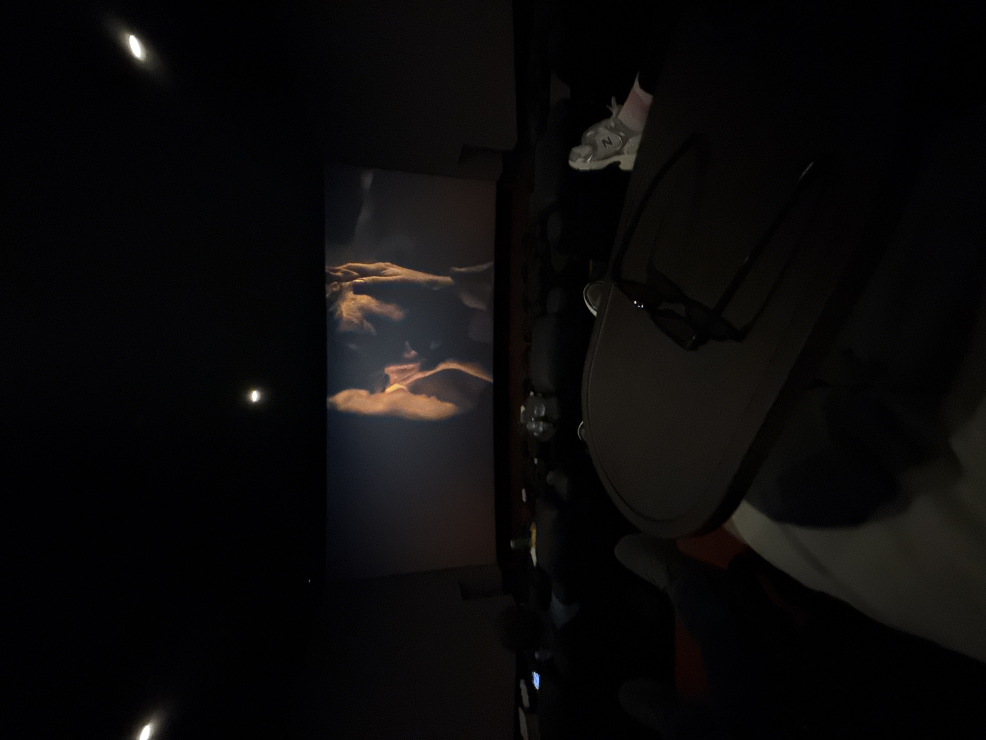 The image depicts the interior of a darkened movie theater, viewed from a seat in a middle row. A large projection screen dominates the front, displaying a film scene in warm, amber tones. The screen content features a close-up of a person with hands near their face, exhibiting an expression that appears distressed or contemplative, while another figure's profile is partially visible to the left.
Above the screen, two recessed ceiling lights emit small, circular beams of light. In the foreground, a dark, round-edged table or tray extends from the viewer's immediate left, on which a pair of dark-framed 3D glasses rests, angled towards the right. To the viewer's immediate right, on an adjacent seat, a pair of light-colored athletic shoes (sneakers) are placed, one upright and one on its side, next to a pink garment.
Several rows of dark, upholstered cinema seats are visible, occupied by an audience whose silhouettes are discernable. Some patrons have food and drink containers on adjacent tables or armrests, with one item appearing as a yellow-capped bottle. One individual in the middle distance has an illuminated smartphone screen visible in their lap. The feet of the person taking the photograph are visible resting on a footrest in the lower left of the frame.
No specific visual identifiers within the image directly indicate the location as Plovdiv, Bulgaria.