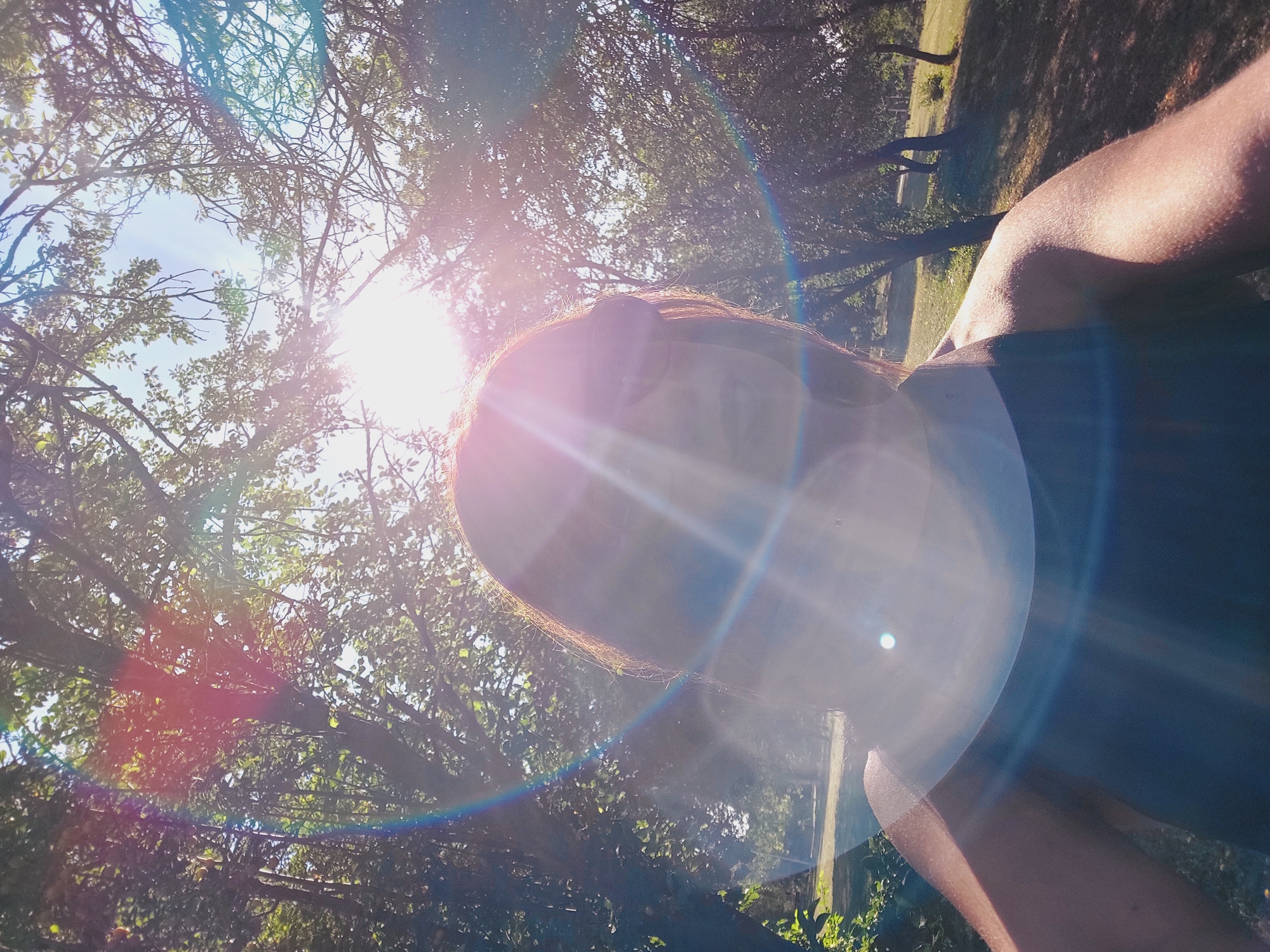 A selfie is taken from a low angle, looking upwards. The sun is directly in front of the camera, causing significant lens flare and overexposure, obscuring the face and parts of the upper body. The person is wearing a dark colored tank top. They have brown hair that is slightly lit by the sun. The background shows a park-like setting with trees and green grass, illuminated by the bright sunlight. The scene appears to be outdoors in a natural environment during the daytime.