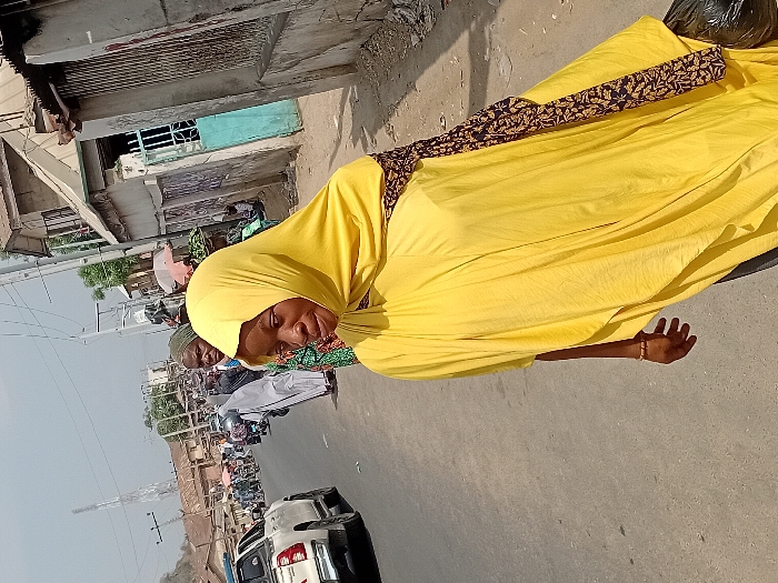 A woman in a yellow hijab and dress walks alongside a busy street. The dress has a dark brown and gold pattern on the upper back. Behind her, a line of cars moves along the road. Buildings line the left side of the frame, with power lines overhead. Other people are also visible in the background.