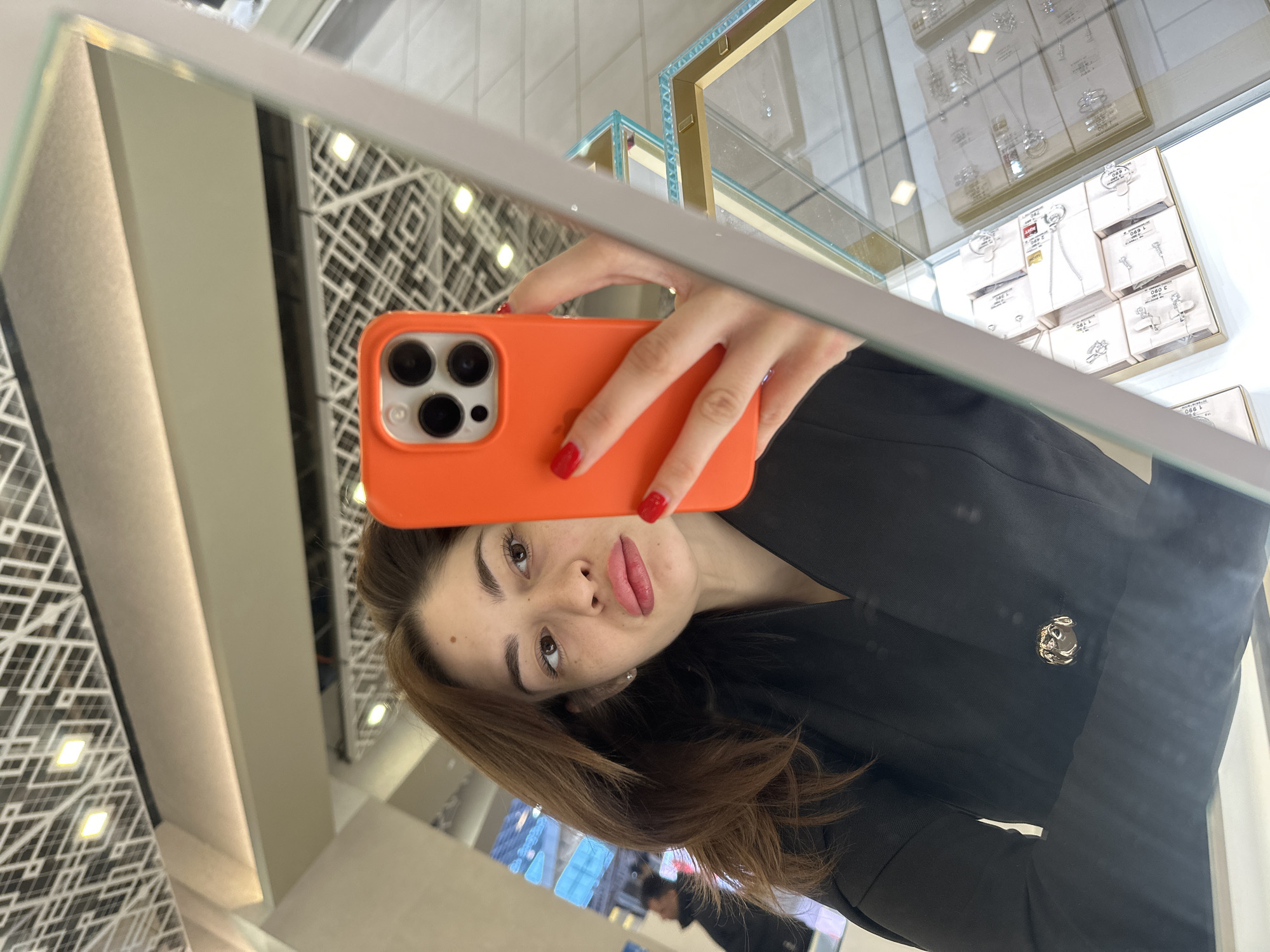 A female individual is depicted in a mirror selfie, captured in what appears to be a SOKOLOV jewelry store located in Yekaterinburg, Russia. The subject, viewed from the chest up, has brown hair, visible freckles on her face, and notably full lips. She wears a black blazer adorned with a prominent gold-colored spherical button on the lapel area. Her right hand, with red-painted fingernails, holds an orange-cased smartphone (exhibiting a triple-camera array) in front of her face, presumably used to take the photograph.
The environment, as reflected in the mirror, features multiple glass display cases positioned to the right of the individual, containing various jewelry items such as necklaces and rings presented on white pads. Price tags displaying Cyrillic text, including "РУБ." (Rubles) and the brand name "SOKOLOV," are discernible within these cases. The display units incorporate gold-toned framing and light blue trim elements. The ceiling, reflected above the subject, showcases a contemporary geometric grid pattern integrated with square recessed light fixtures. A blurred reflection of another person or object is visible in the background to the left of the main subject.