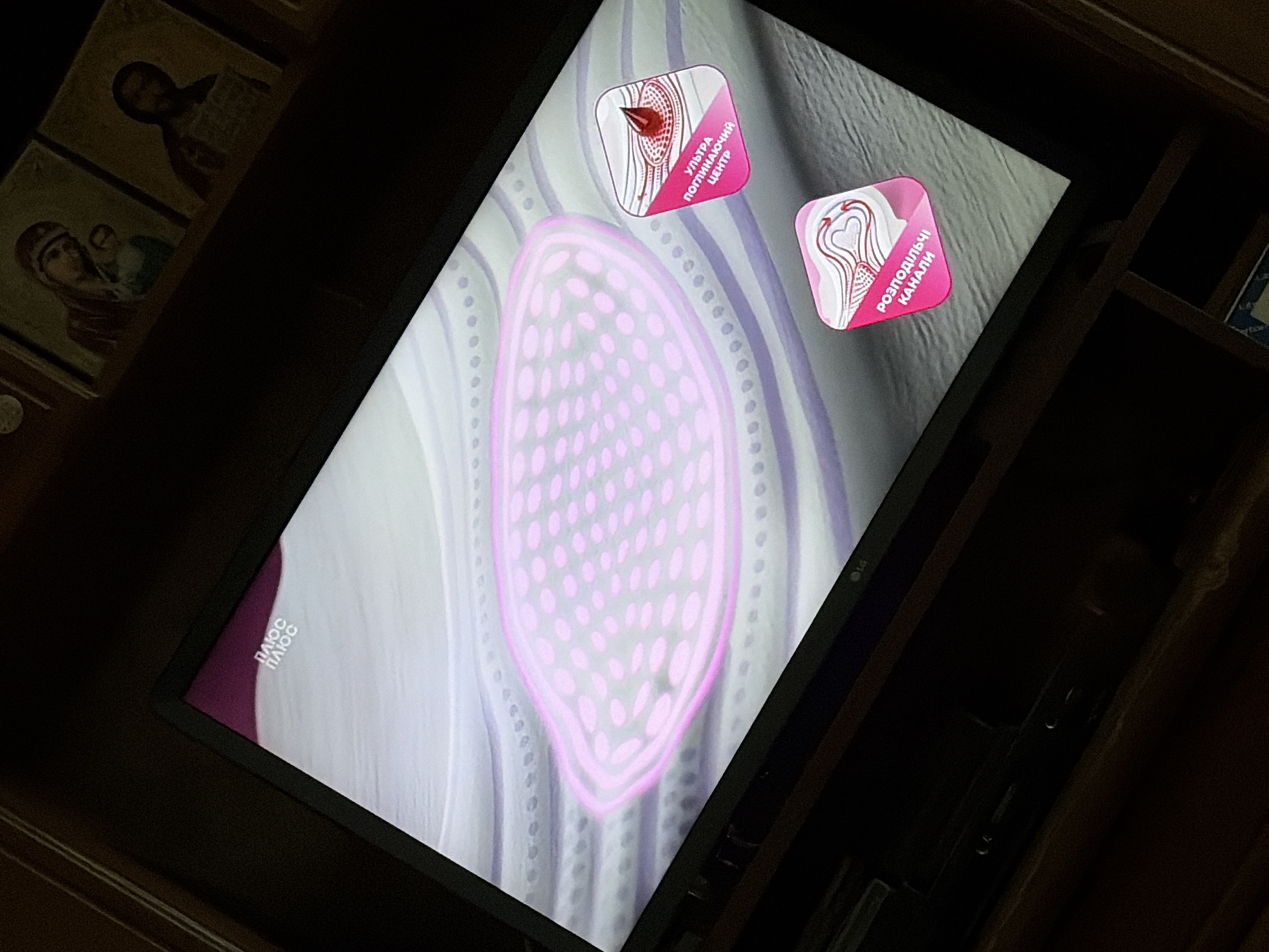 The image shows a television screen displaying a graphic. The graphic features a large, light pink, oval shape with a textured interior of smaller pink ovals. Surrounding this central shape are wavy, light purple and white stripes. In the lower right portion of the screen, there are two smaller, pink, square-shaped graphics with rounded corners. The top graphic contains a red droplet within a white area, and text in a dark color that reads "УЛЬТРА ПОГЛИНАЮЧИЙ ЦЕНТР". The bottom graphic displays a heart-shaped pattern with arrows, and text that reads "РОЗПОДІЛЬЧІ КАНАЛИ". In the upper left corner of the screen, a logo with the text "ПЛЮС ПЛЮС" is visible. The television itself is dark, with a visible "LG" logo at the bottom center of the screen. The background behind the television is dark, with some illuminated religious icons visible on a shelf.