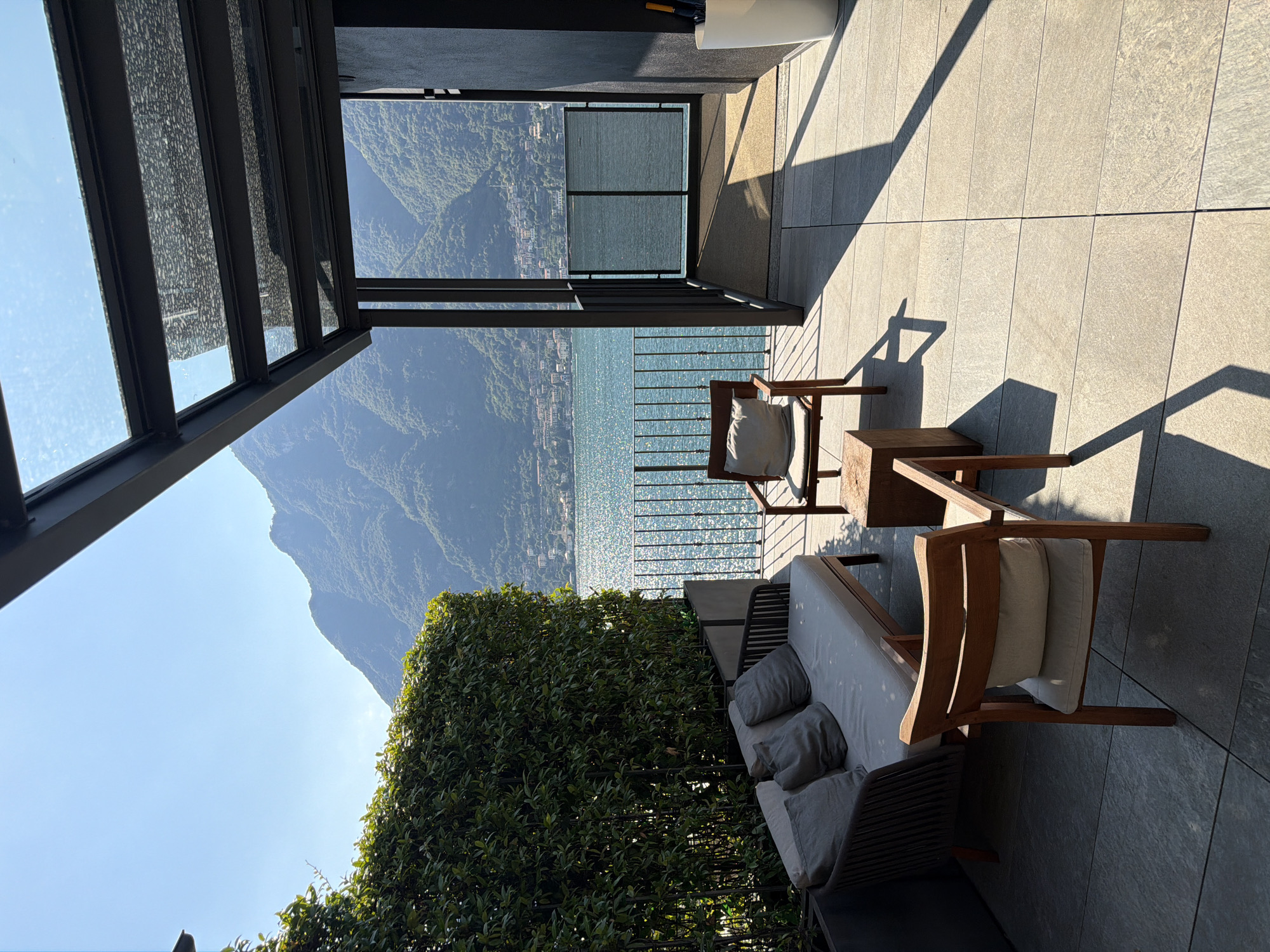 A sunny day is depicted on a balcony overlooking a lake and mountains in Pognana Lario, Italy. The balcony is furnished with a modern sofa and two wooden chairs with light-colored cushions, and a small wooden table between them. A black metal and glass canopy extends over part of the balcony. A tall green hedge lines the left side of the balcony, and a black metal railing separates the balcony from the view. The lake, with its surface sparkling in the sun, is visible beyond the railing, as are lush green mountains in the distance. A dark wall is on the right side of the balcony, with a white planter containing an umbrella. The floor is tiled with large, light-colored square tiles.