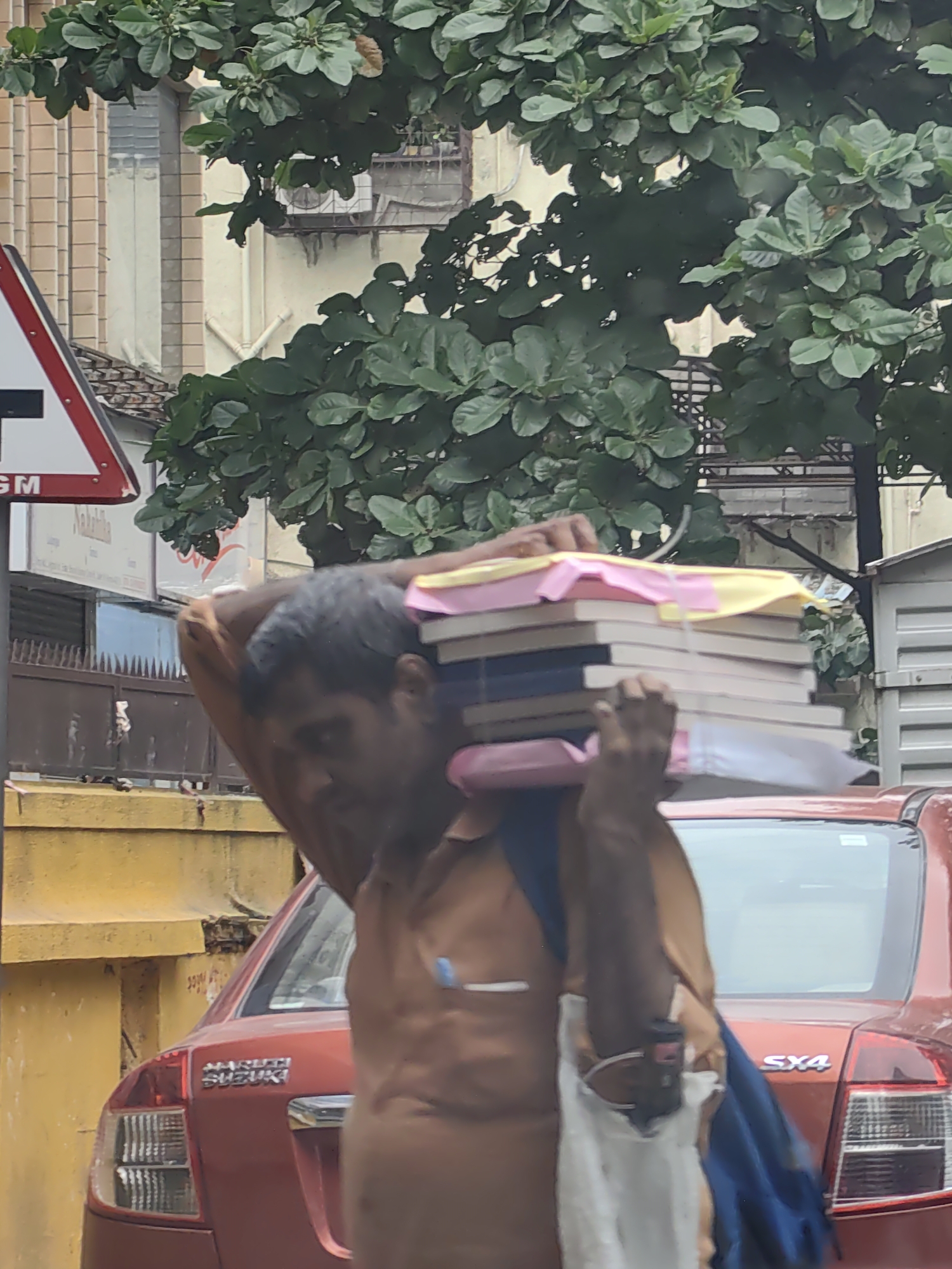 A man walks through a city street in Mumbai, India, carrying a stack of books on his shoulders. The books are topped with yellow and pink paper. He is wearing a brown shirt and has a blue backpack slung over his shoulder. Behind him, a red Maruti Suzuki SX4 car is parked. The background shows a yellow wall, a traffic sign with a red border and a white center with a black "T" shape, and lush green trees with dense foliage. A building with a tiled facade is visible behind the trees. The scene appears to be an outdoor urban environment, possibly during the daytime.