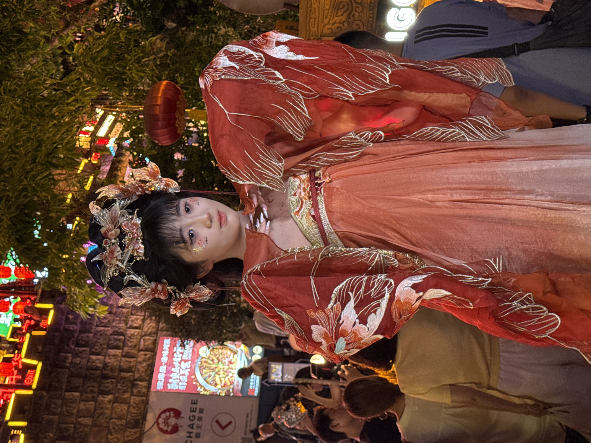 The image captures a nighttime street scene in Yuzhong District, China, possibly during a festival or event, as indicated by the abundant colorful lighting and decorative lanterns. A young woman, likely a performer or participant, is the central subject, dressed in an elaborate traditional Chinese costume. Her attire includes a layered, flowing orange dress and a dramatic red cape adorned with white floral and leaf embroidery. She wears an ornate headdress with floral and butterfly motifs, and her makeup features delicate glitter accents on her cheeks.
She is positioned in the foreground, looking directly at the viewer with a gentle expression. Behind her, a crowd of people is visible, some holding up phones, suggesting they are taking pictures or videos. The background is a blur of activity and illumination, with neon signs, brick walls, and lush green foliage creating a vibrant, festive atmosphere. A prominent red lantern hangs from a tree in the middle ground. The overall scene conveys a sense of celebration and cultural immersion within a bustling urban environment.