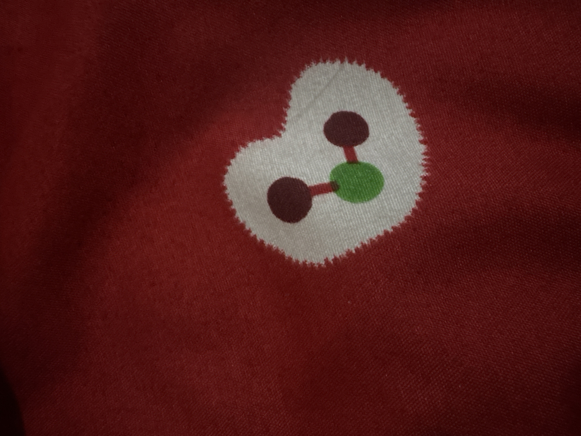 The image displays a close-up view of a red fabric with a white heart-shaped design featuring a stylized molecular structure. The structure consists of three circles connected by lines. The central circle is green, and the two outer circles are dark red. The heart shape has a serrated edge. The red fabric has a visible texture. The image does not contain any information about the city of Danawinangun, Indonesia, nor does it depict any people, actions, or interactions.