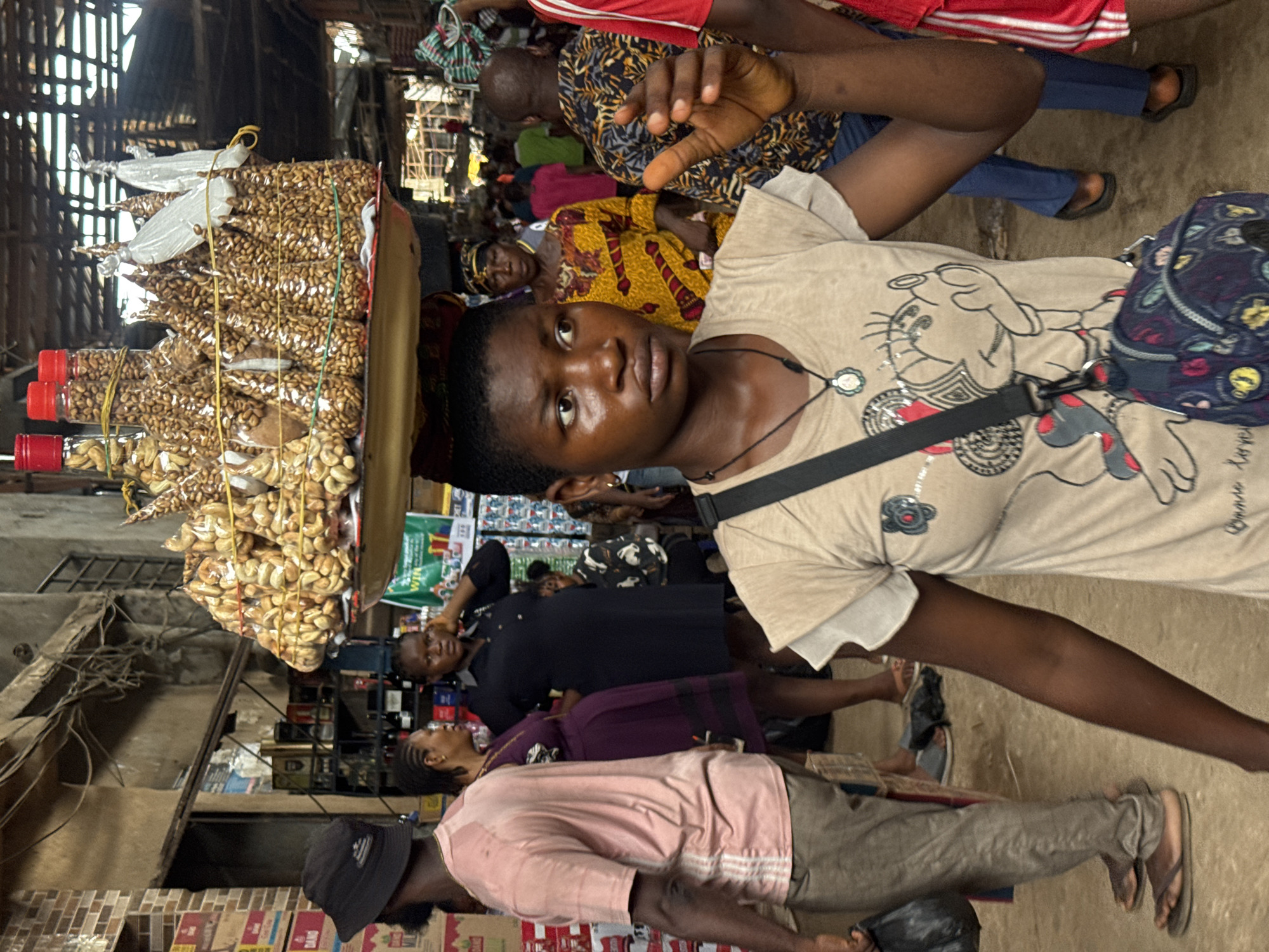 The image depicts a bustling market scene in Onitsha, Nigeria. A young woman, likely a vendor, carries a large tray laden with packaged goods, possibly nuts or seeds, on her head. She is in the foreground, looking directly at the camera with a serious expression. She wears a light-colored t-shirt with a cartoon graphic and a dark crossbody bag. The market scene is crowded with people, many of whom are in motion. Several individuals are discernible, including a woman in a yellow patterned dress, a man in a red and white striped shirt, and other shoppers and vendors. The background reveals market stalls and merchandise. The lighting suggests an indoor market space, possibly with a roof overhead. The overall impression is of a vibrant, busy marketplace.