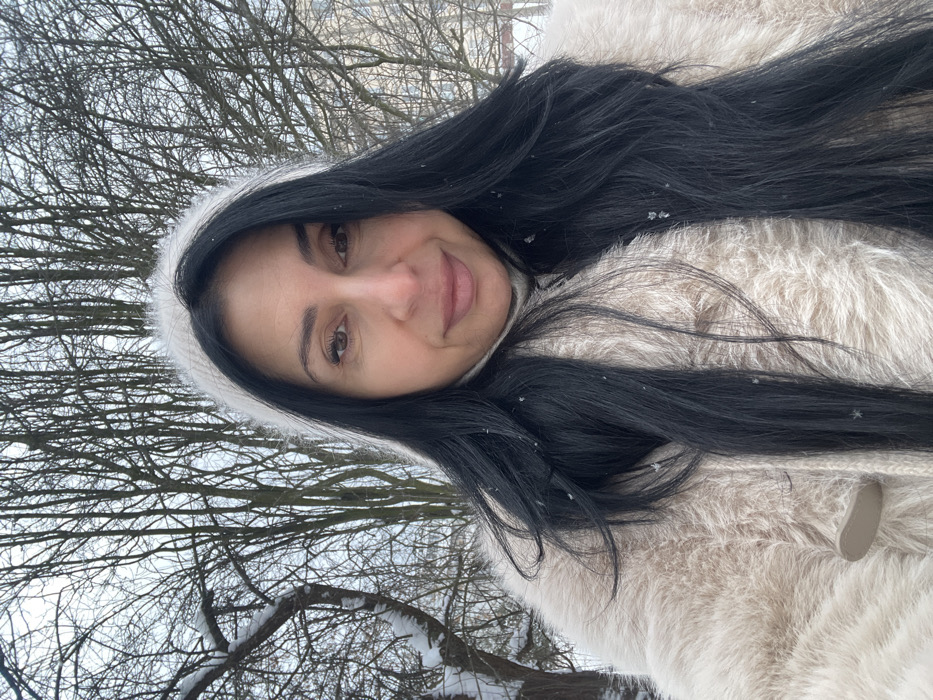 The image depicts an adult female outdoors in a snowy, wooded environment within the city of Syasstroy, Russia. The woman, positioned centrally and looking directly at the camera, has long, dark hair, a medium skin tone, and a subtle smile. She is dressed in a light cream or beige long-haired faux fur coat, exhibiting a visible light-colored toggle closure on the left side, over what appears to be a light-colored turtleneck. A fuzzy, light-colored headband or hat portion is visible, framing the top of her dark hair. Small, distinct snowflakes are scattered on her hair and coat. The background features numerous bare deciduous trees, their branches lightly dusted with snow, under an overcast sky. A light-colored building is faintly discernible through the trees in the upper right background. The scene conveys a cold, winter setting with active or recent light snowfall.